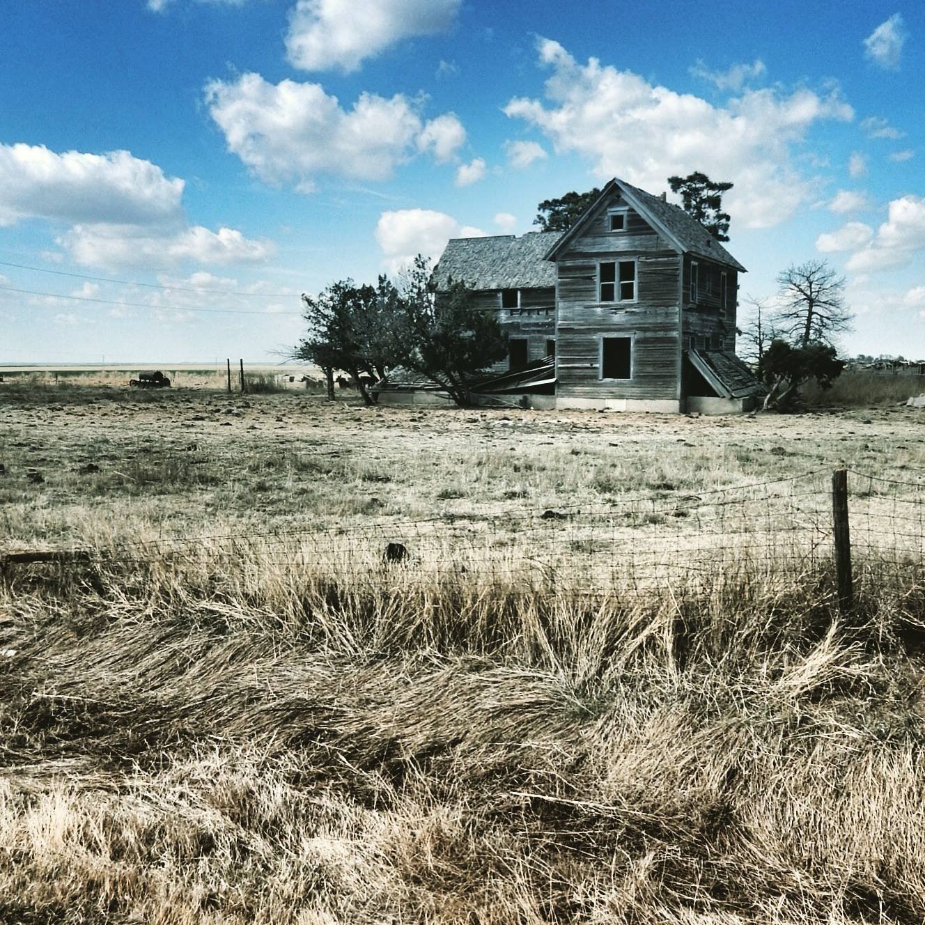 An abandoned, dilapidated wooden house in a grassy field under a partly cloudy sky.