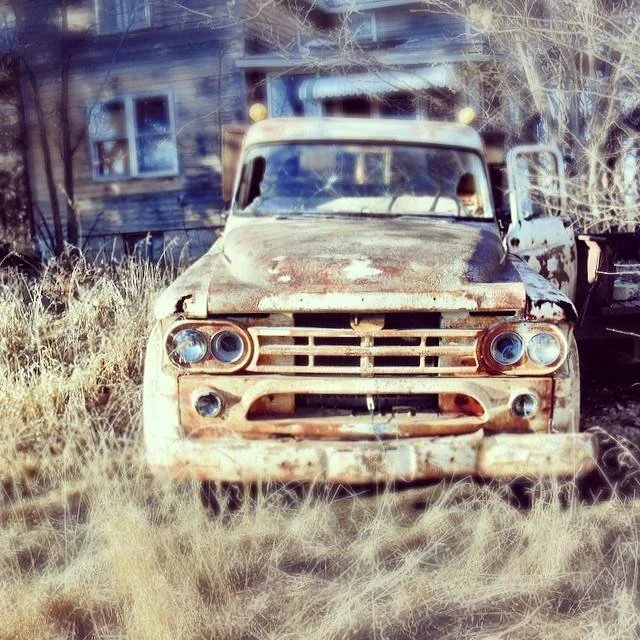 Old, rusty vintage truck abandoned in a grassy yard with a weathered house in the background.