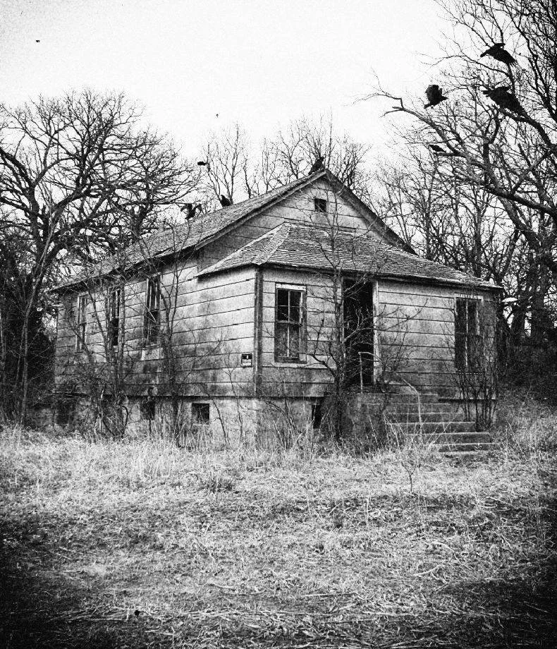 An old, abandoned house with broken windows and peeling paint, surrounded by leafless trees and overgrown grass, under an overcast sky.