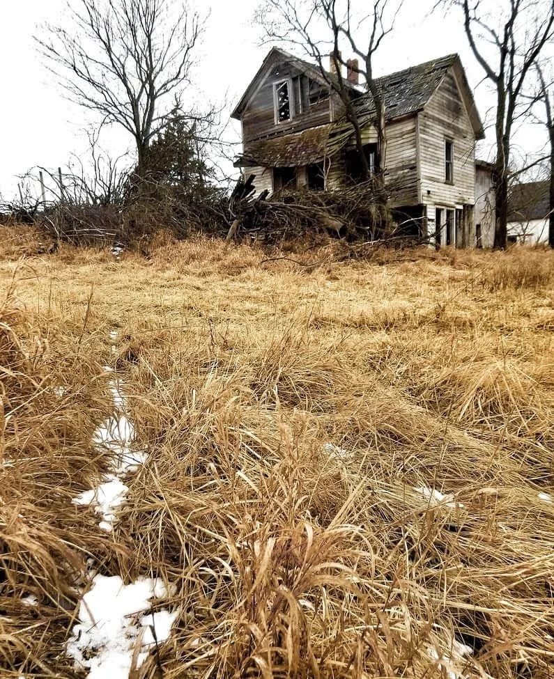 An abandoned, dilapidated house with broken windows and rotting wood, surrounded by leafless trees, with dry grass and patches of snow in the foreground.