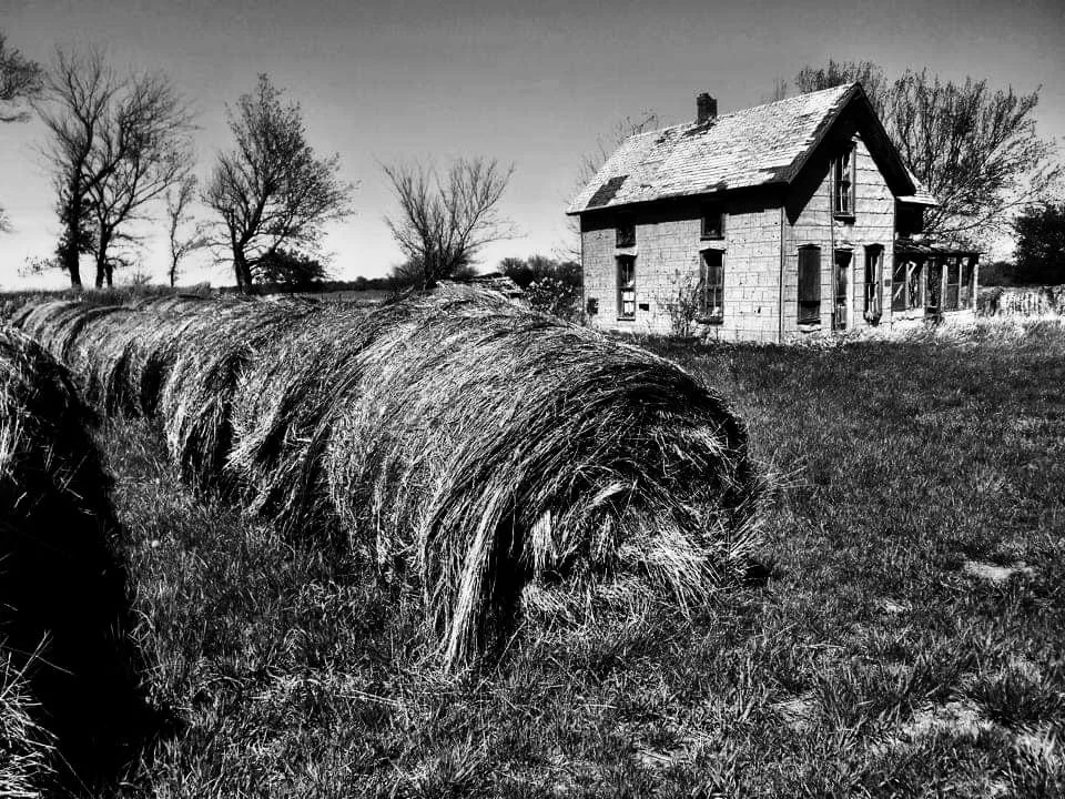 An old, abandoned house with boarded-up windows in a rural area. In the foreground, there is a large hay bale on the grass. Bare trees are visible in the background, and the sky appears cloudy. The photo is in black and white.