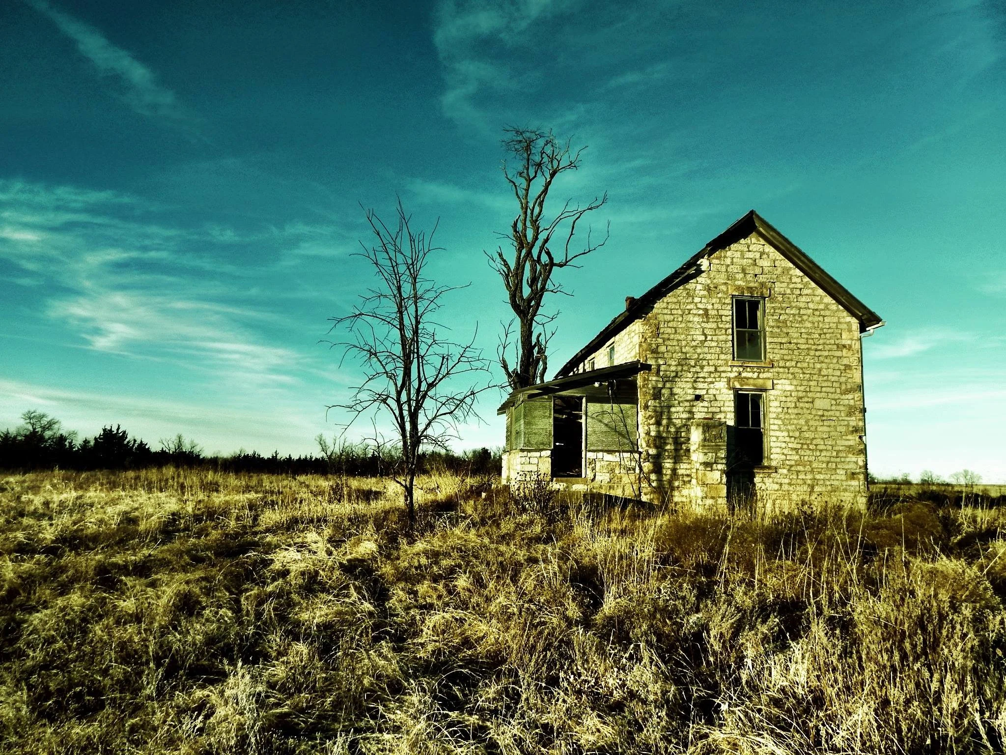 An abandoned, dilapidated stone house with broken windows sits in a field of dry grass. Two leafless trees are beside the house under a blue sky with scattered clouds.