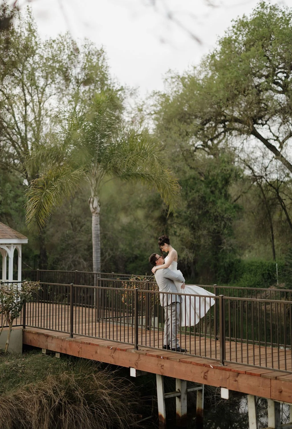 A small white gazebo with a shingled roof in a lush green garden with tall trees and palm trees.