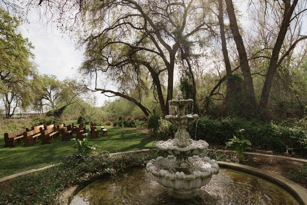A bride and groom embrace outdoors during their wedding, surrounded by floral decorations, with trees and a grassy area in the background.