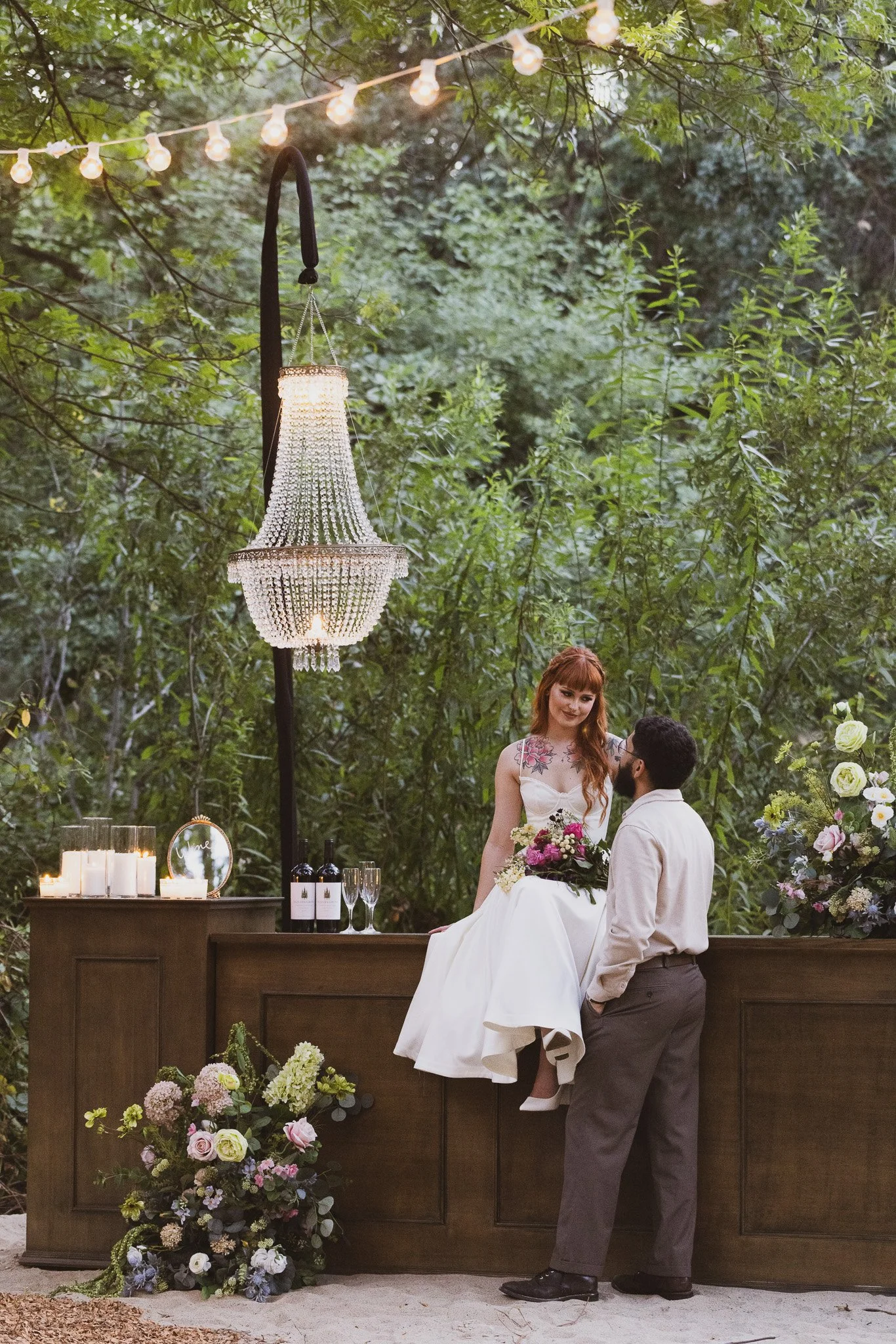 A bride and groom in a wedding scene outdoors, with the bride sitting on a wooden table holding a bouquet of pink and white flowers, and the groom standing in front of her. The scene is decorated with candles, flowers, and a chandelier hanging from t