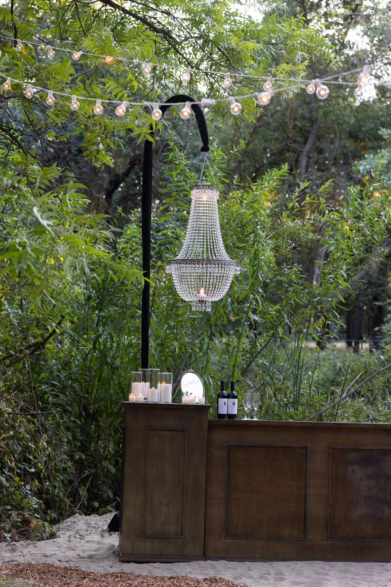 Outdoor setting with a wooden bar, candles, two wine bottles, two wine glasses, a mirror, a chandelier, string lights, and lush green trees in the background.