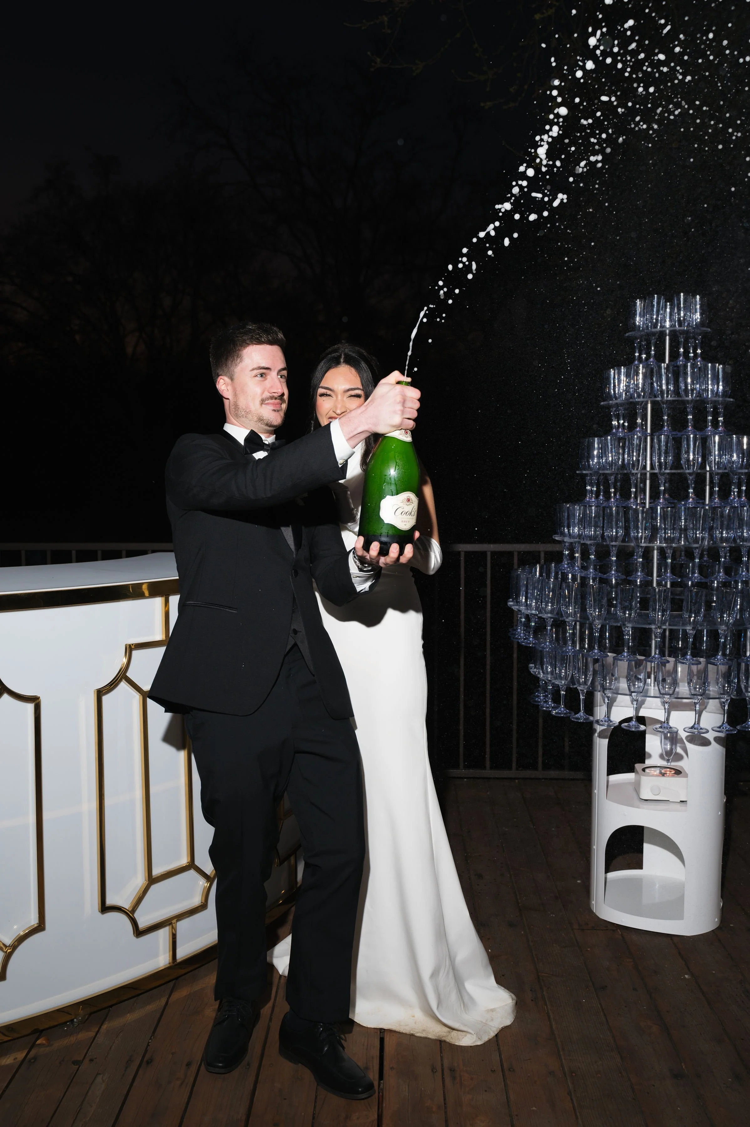 A couple in formal attire celebrates by popping a champagne bottle on a roof deck at night, with a tower of empty glasses in the background.