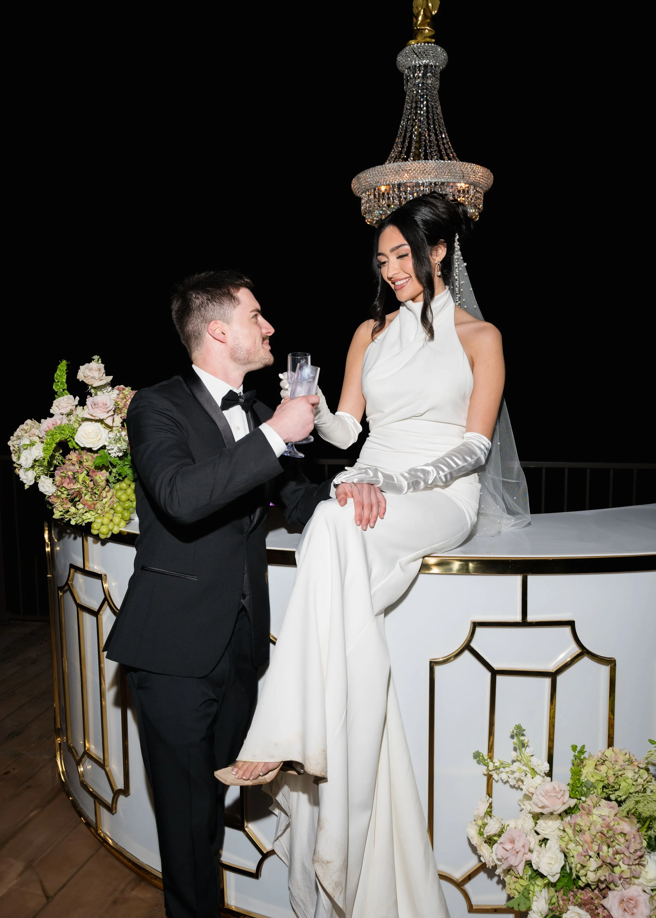 A couple at a wedding celebration, with the woman wearing a white dress and sitting on a bar counter, and the man in a tuxedo offering her a drink. There is a chandelier above them and floral arrangements nearby.