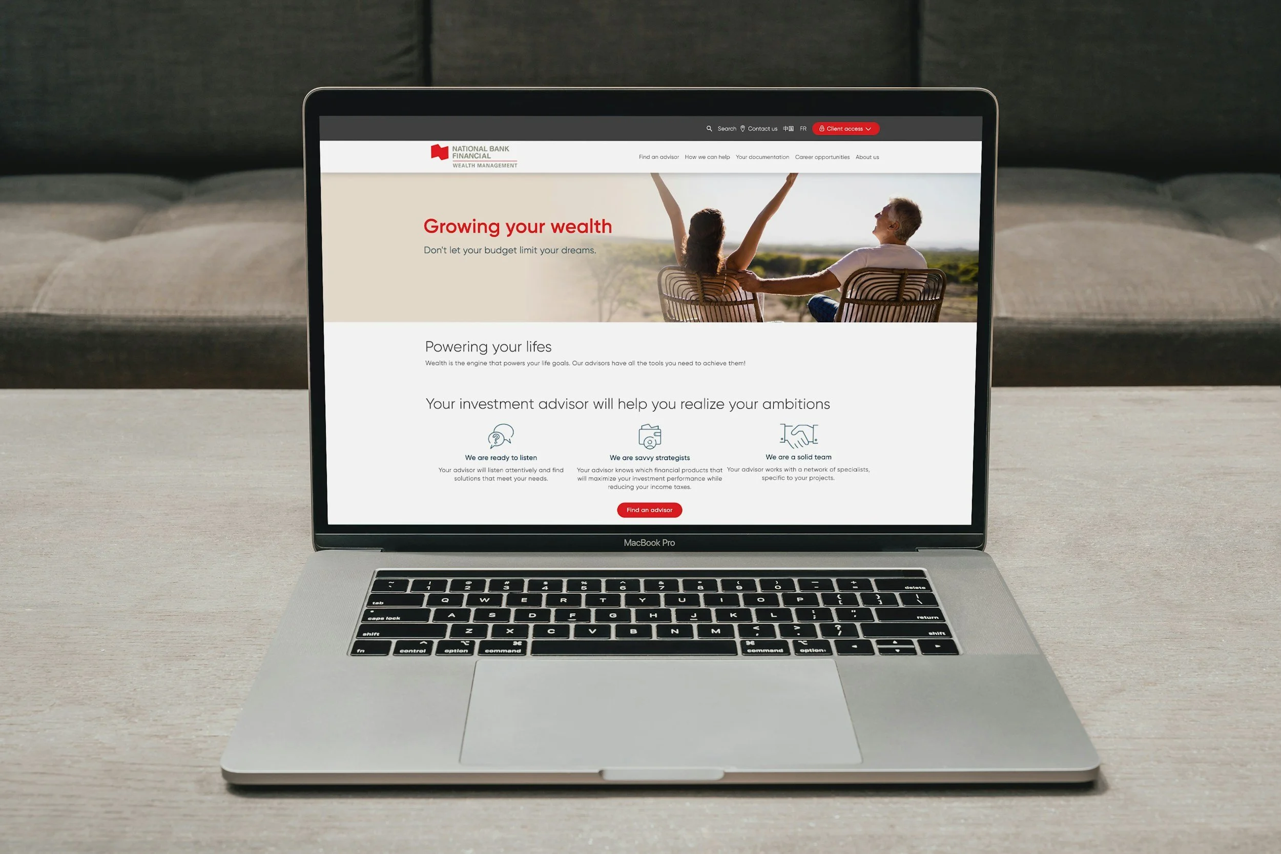 Open MacBook Pro laptop on a desk displaying a financial services website with the headline "Growing your wealth" and an image of two people sitting outside, one of whom has arms raised, enjoying the view.