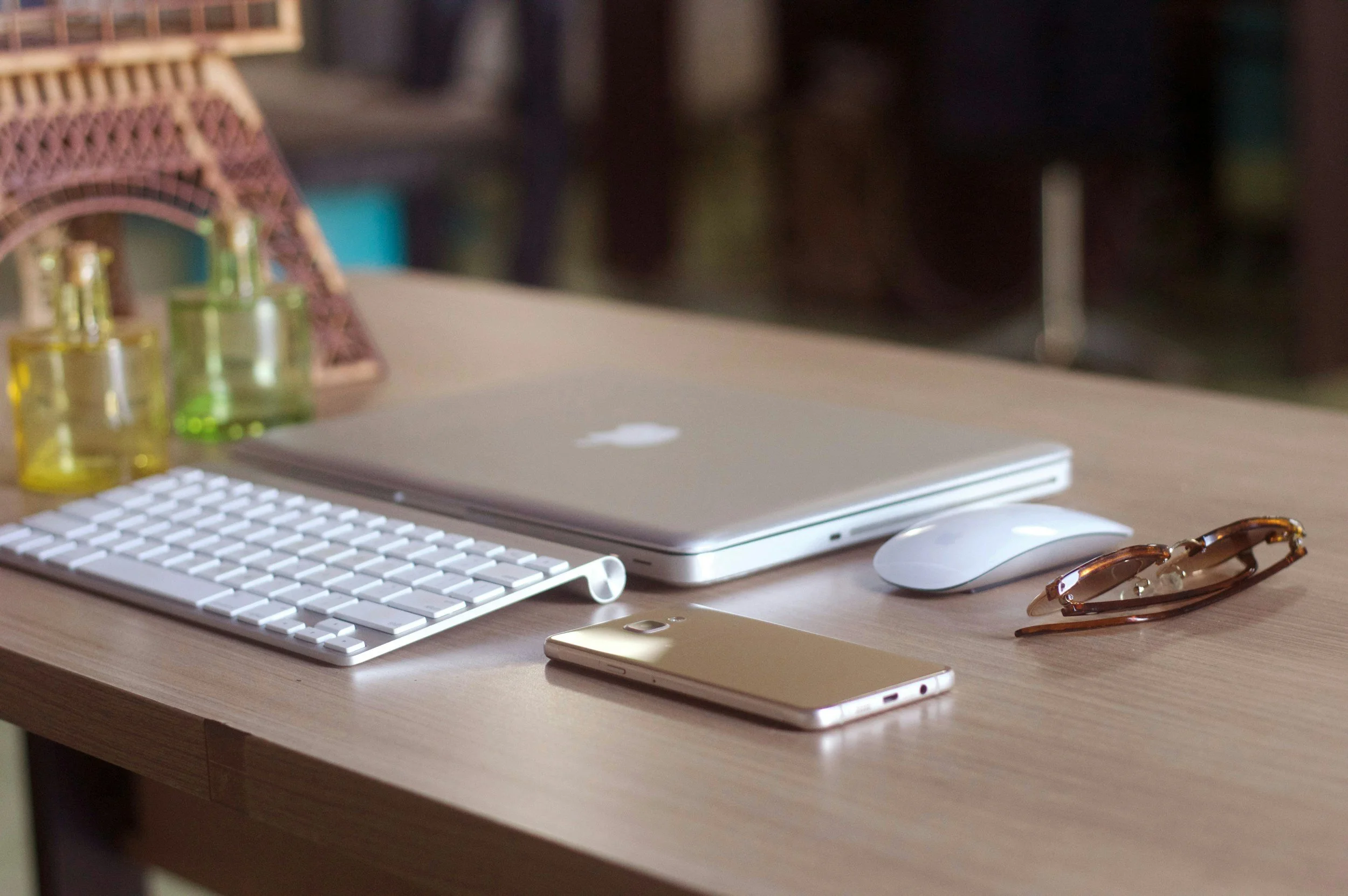 A wooden desk with a closed Apple MacBook, a white wireless mouse, a gold smartphone, a white wireless keyboard, and a pair of sunglasses. In the background, there are three small bottles with different colored liquids and a decorative eiffel tower model.