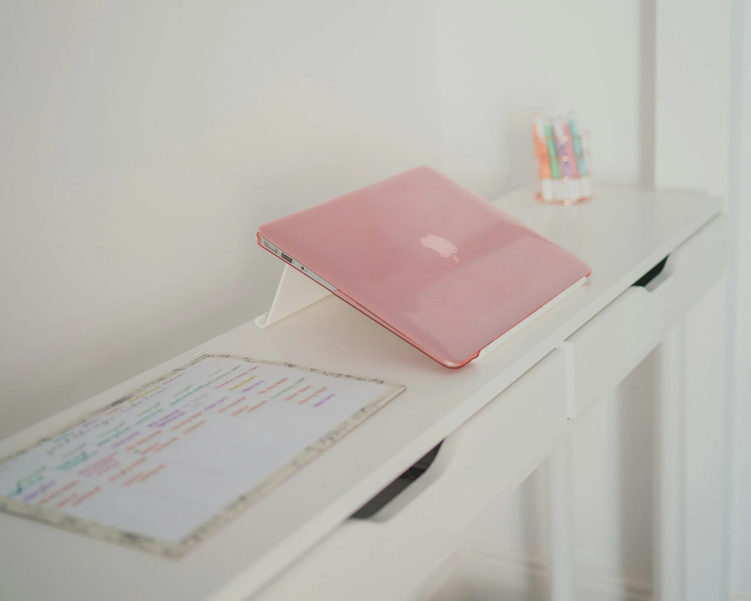 A pink closed MacBook laptop on a white desk, with a colorful handwritten schedule or planner and a small container holding colored pens in the background.
