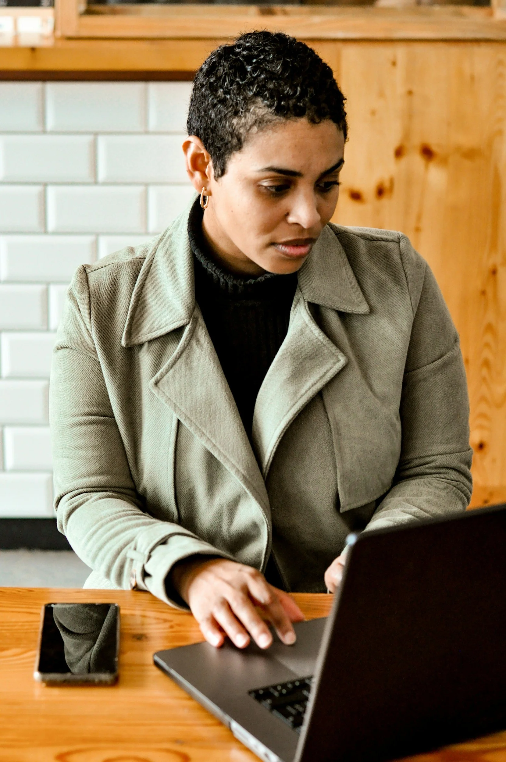 A woman with short, curly hair, wearing a gray jacket and black shirt, working on a laptop at a wooden table in a cozy indoor setting.