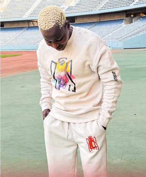 A person with bleached blonde hair wearing a white sweatshirt and white pants standing on a sports field.