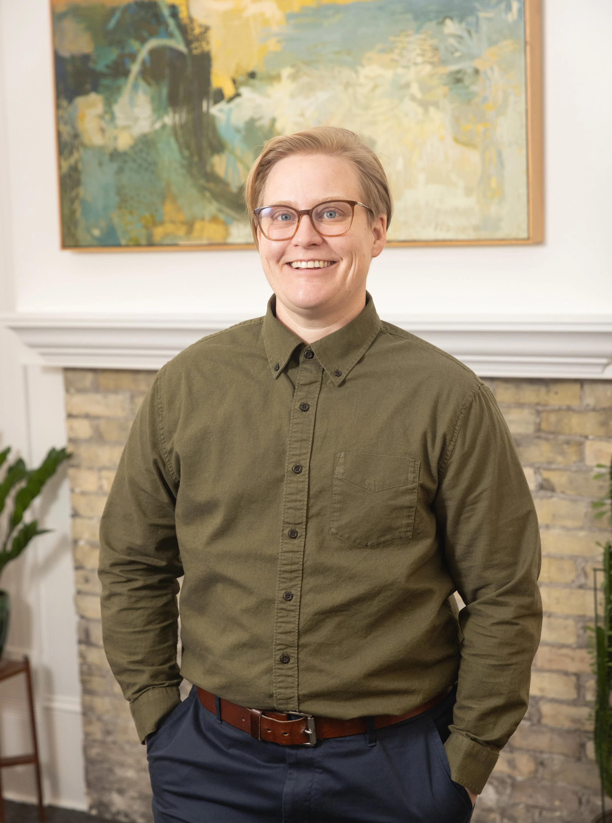 A smiling person with short hair, glasses, wearing an olive green button-up shirt and navy pants, standing in front of a fireplace with a painting above it.