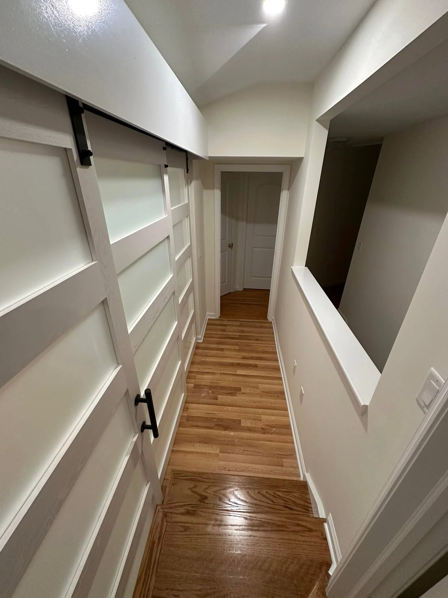 View of a narrow hallway with hardwood flooring, white walls, and a sliding glass door with frosted glass panels on the left. A door with a round doorknob is at the end of the hallway. There is a large rectangular cutout in the wall on the right side