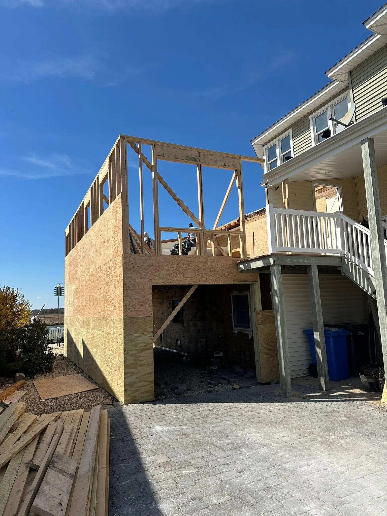 Construction of an outdoor wooden deck attached to a house, with framing in progress and building materials nearby under a clear blue sky.