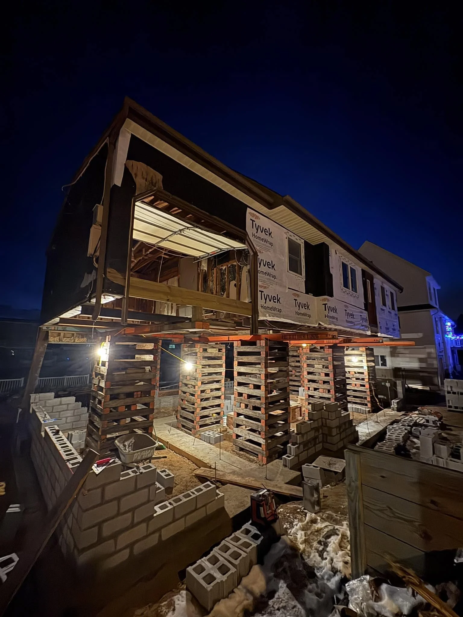 Construction site at night showing a partially built house with framing, insulation, and brickwork, surrounded by building materials and tools.