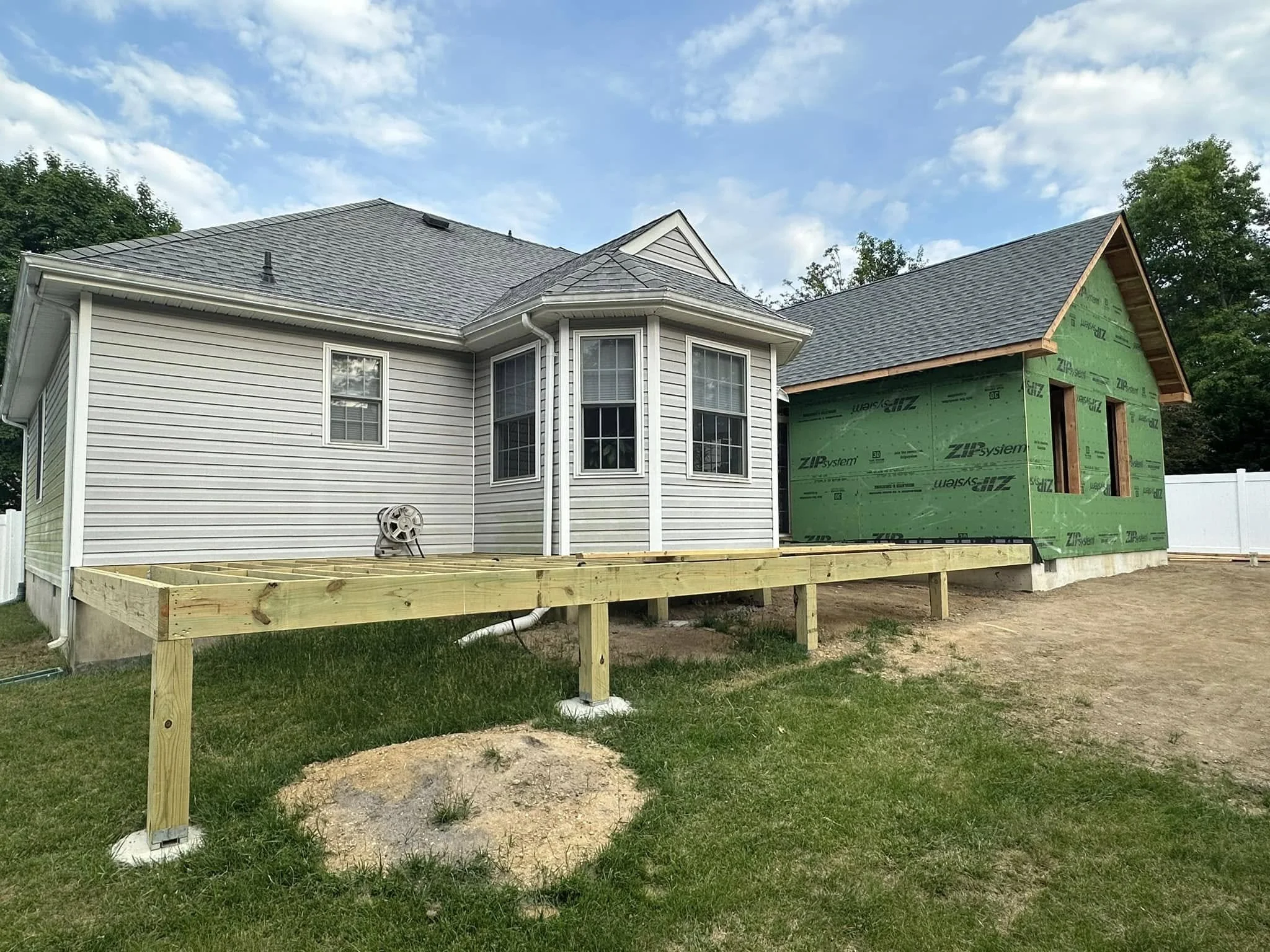 A two-story house under construction with siding and a finished roof. The right side of the house shows green exterior sheathing and open framing. There is a wooden deck frame in the foreground and a cloudy sky above.
