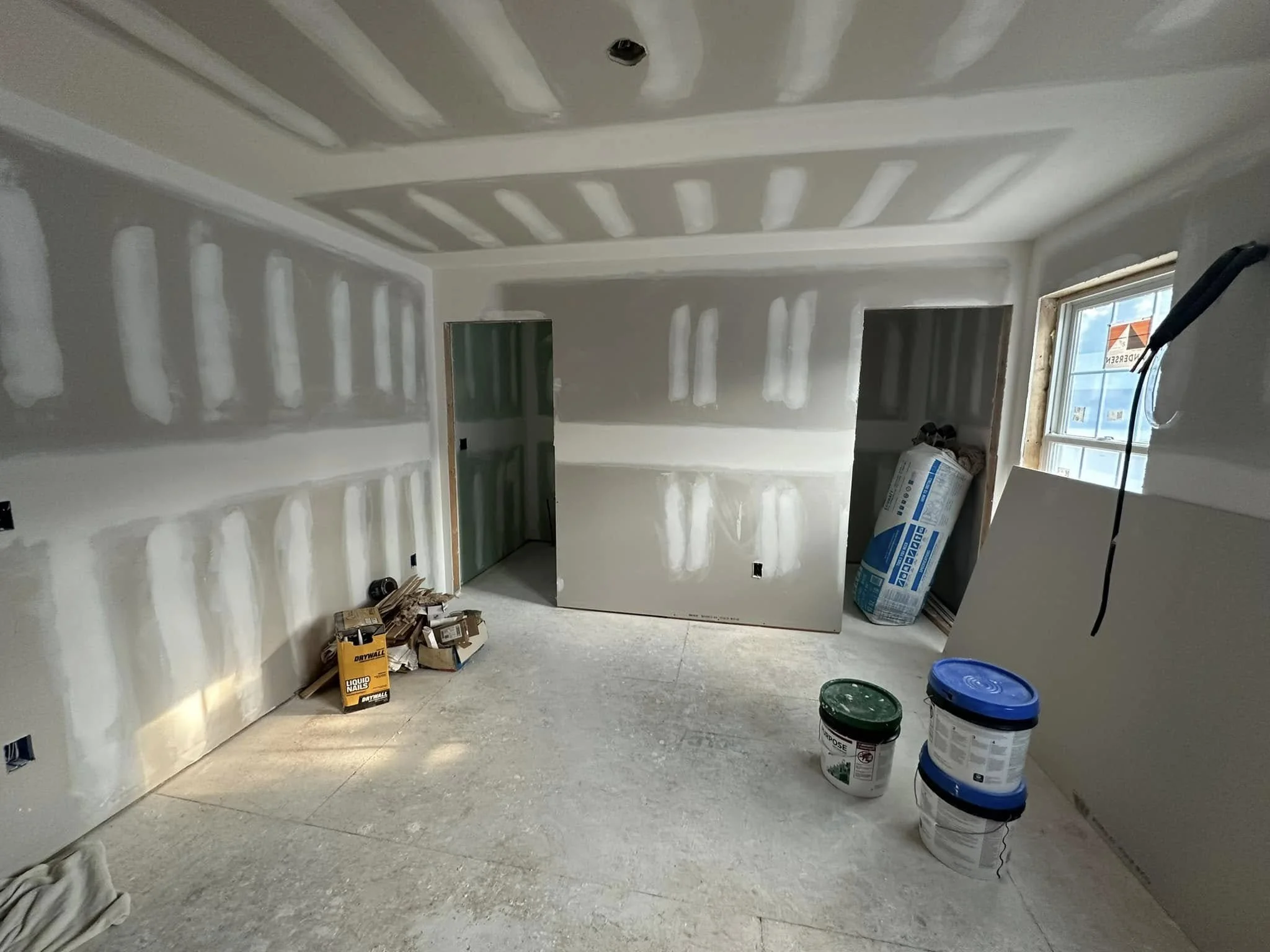 An unfinished room under construction with drywall installed but not yet painted or finished, featuring a ceiling with shadow patterns, a window with sunlight, and construction materials such as paint buckets, drywall, and packaging on the floor.