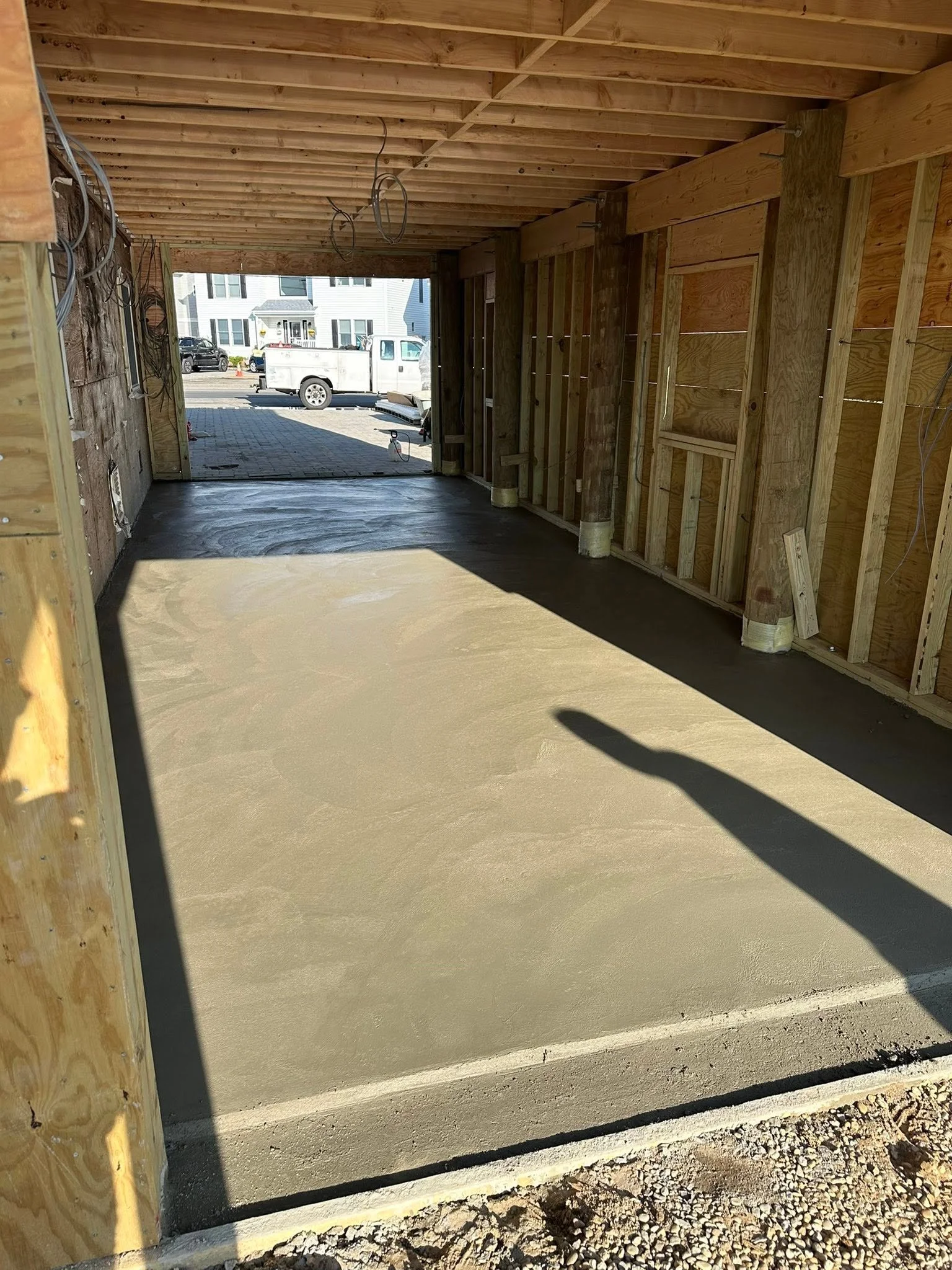 View of a garage with a freshly poured concrete slab floor, unfinished wooden framing, and an open entrance leading to a driveway with parked vehicles and neighboring houses.