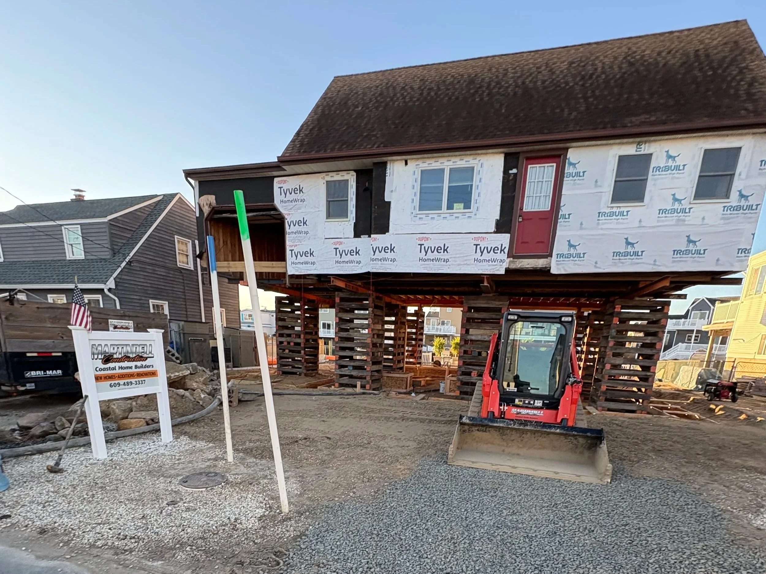 A house under construction elevated on wooden stilts with a small red door, surrounded by construction equipment, gravel, and nearby houses.