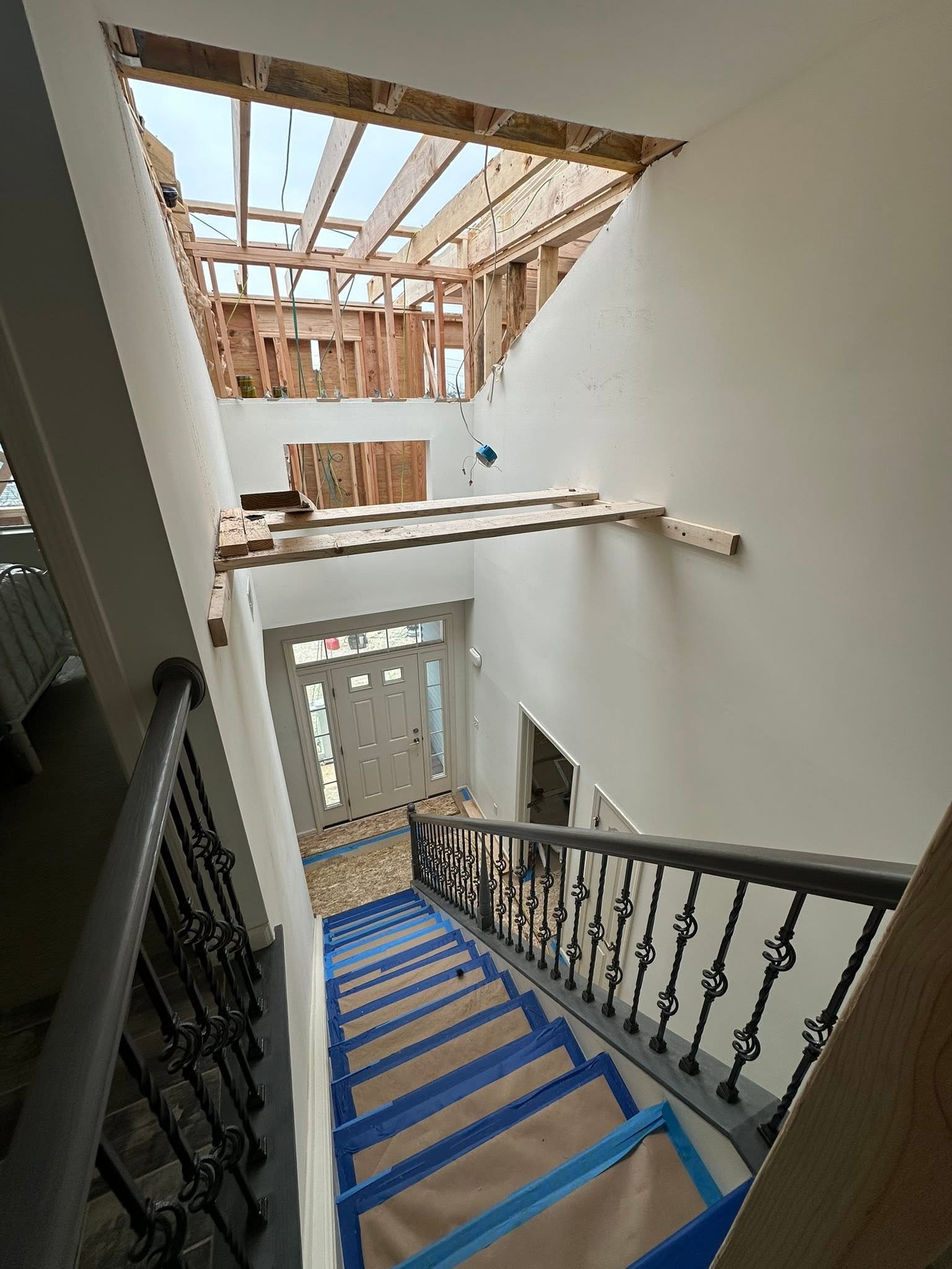 Interior of a house under construction, showing stairs with blue painter's tape and a missing ceiling in the upper level, revealing wooden beams and framing.