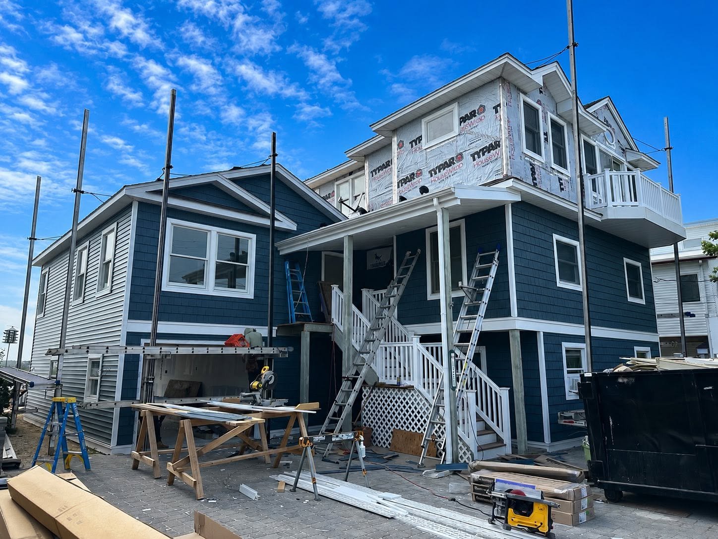 A house under construction with scaffolding and ladders, blue exterior siding, and construction tools and materials scattered around.