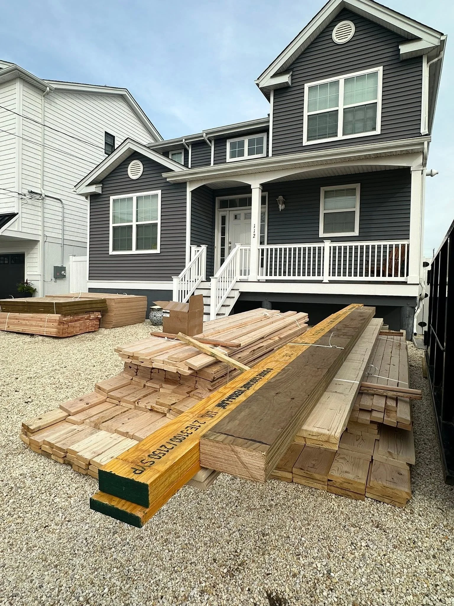 Stacks of wooden planks and lumber on gravel ground in front of a dark blue house under construction.