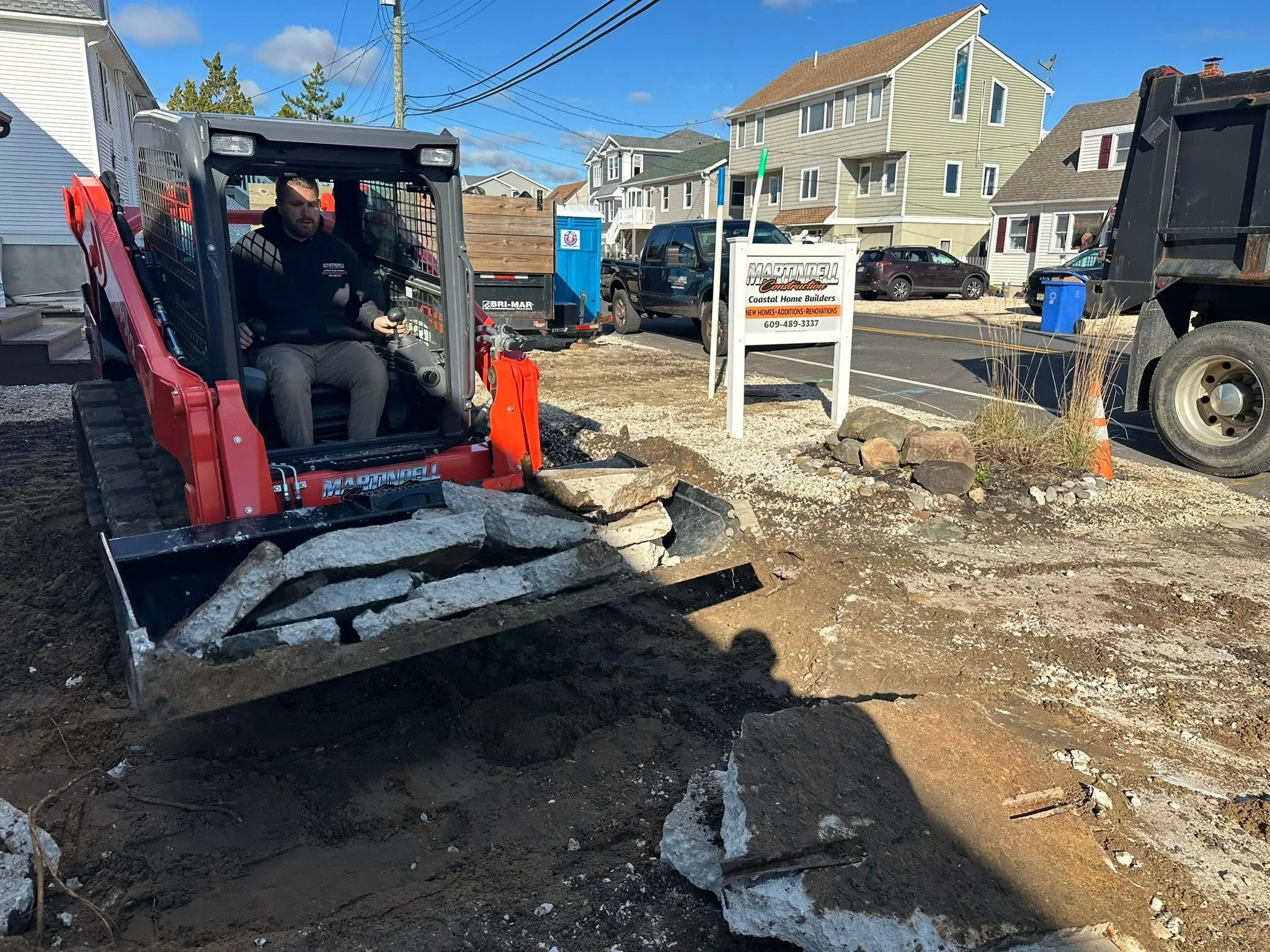 Construction worker operating a mini skid steer loader on a residential street with houses and parked cars, preparing the ground for landscaping.