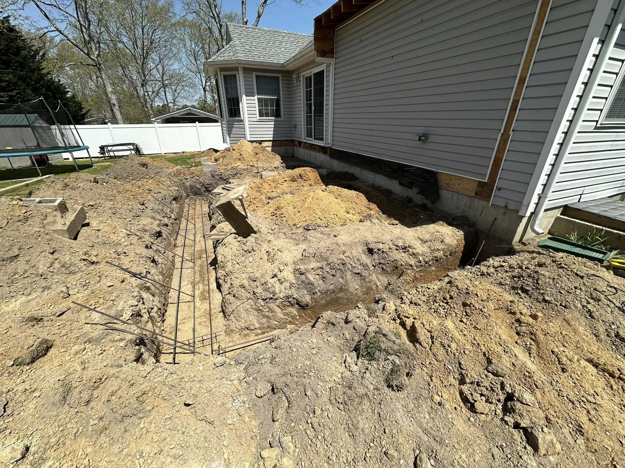 Backyard with construction work in progress, showing excavated dirt and rebar, with a house with siding and windows in the background, along with trees and a sunny sky.