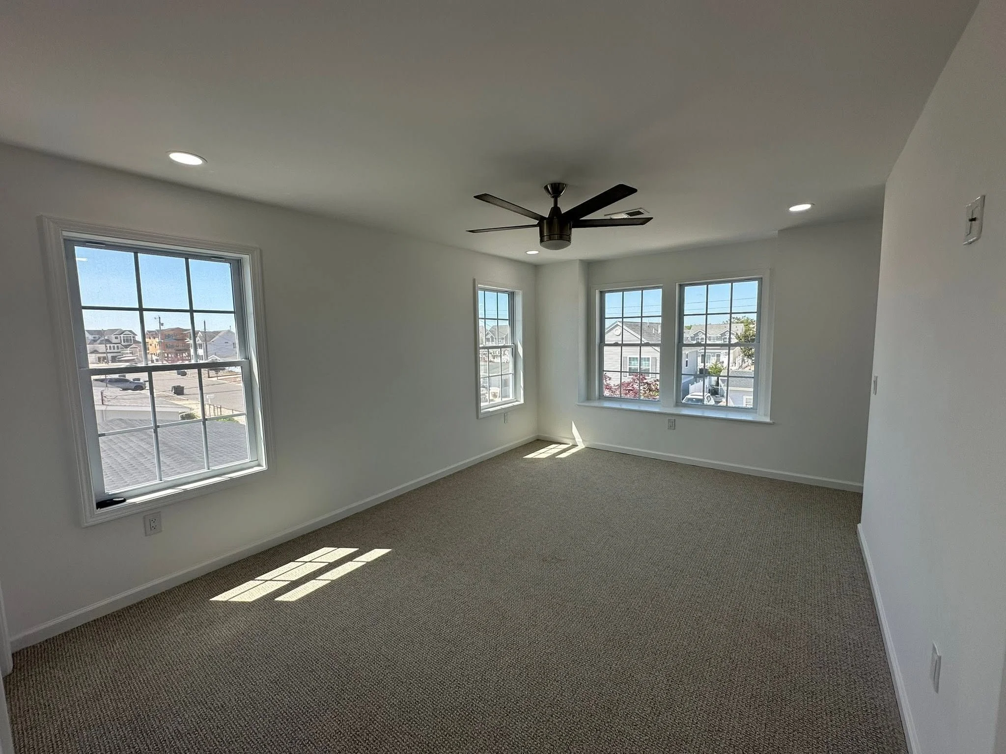A bright and clean corner of a room with three large windows letting in natural light, white walls, a ceiling fan, beige carpet, and ceiling recessed lights.