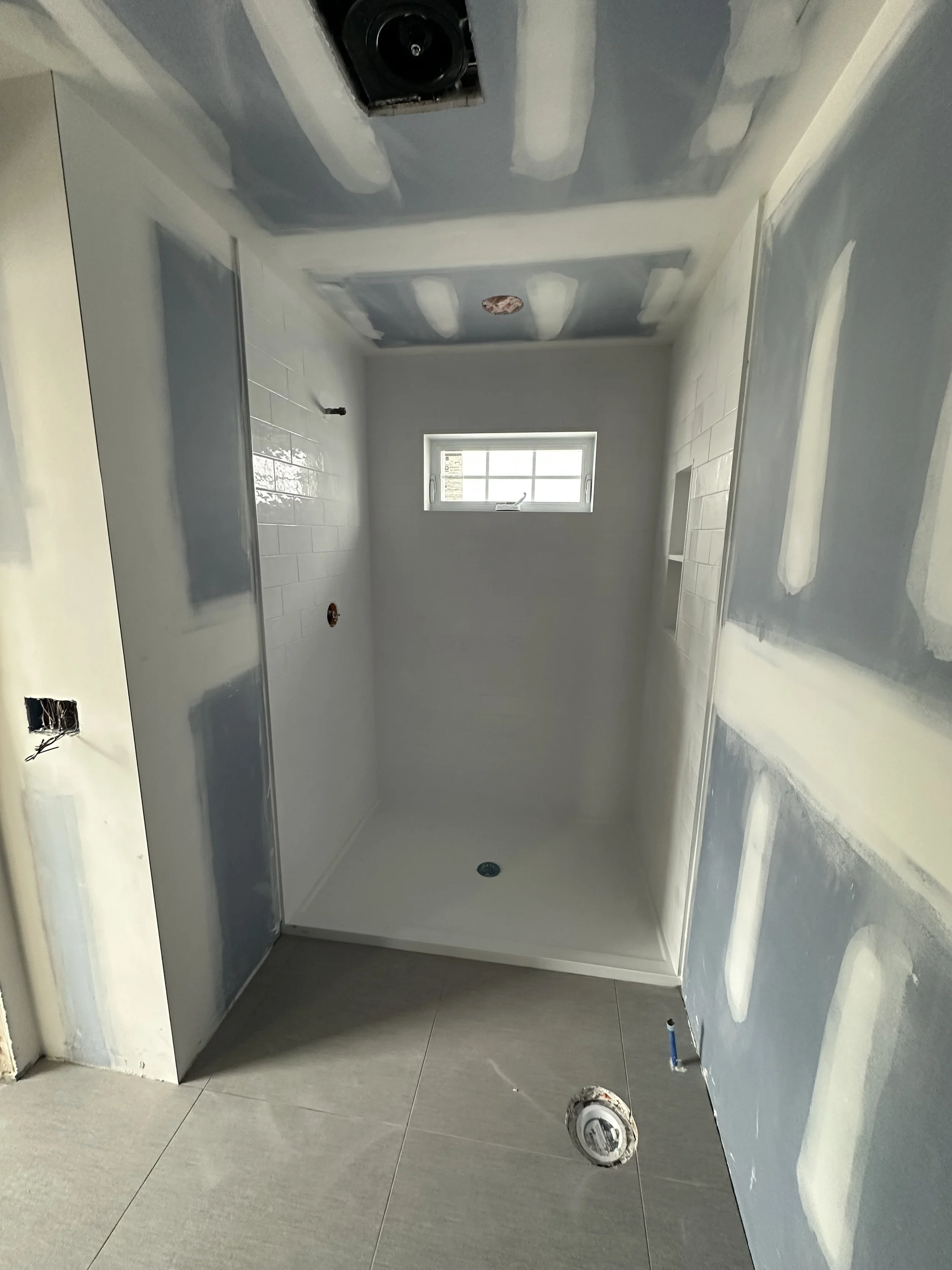 Bathroom with grey tiles and white partial wall with tile molding, window, and fixtures for a shower or bathtub installation under construction.