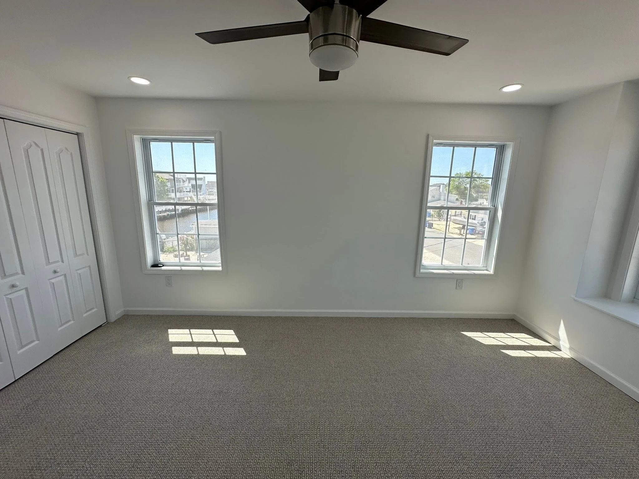 Empty room with white walls, two large windows, beige carpet, white closet doors, and a ceiling fan with dark blades and a frosted glass light cover.