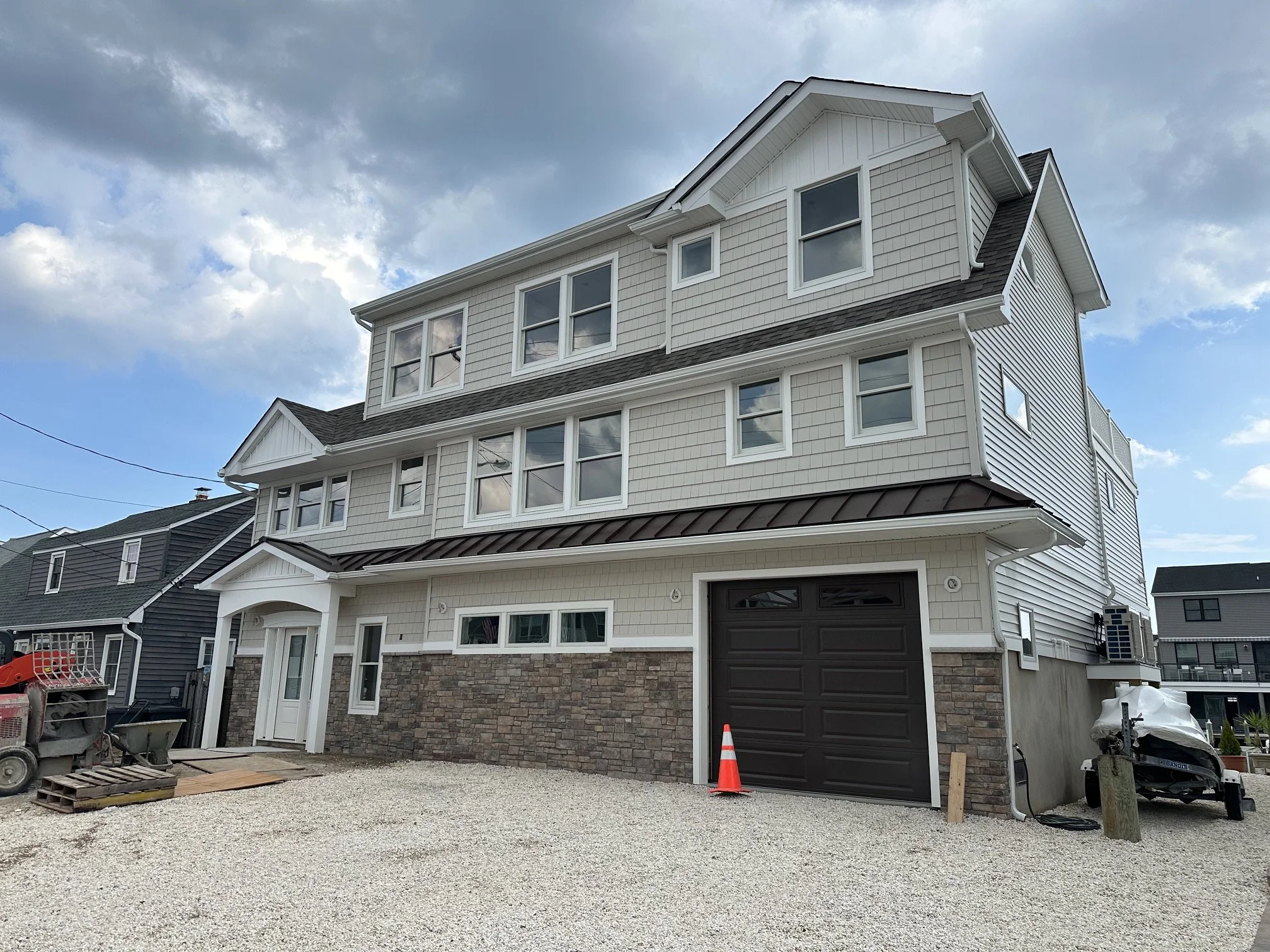 Newly constructed multi-story house with a white exterior, stone basement, and black garage door, under a cloudy sky, with construction equipment and materials at the front.