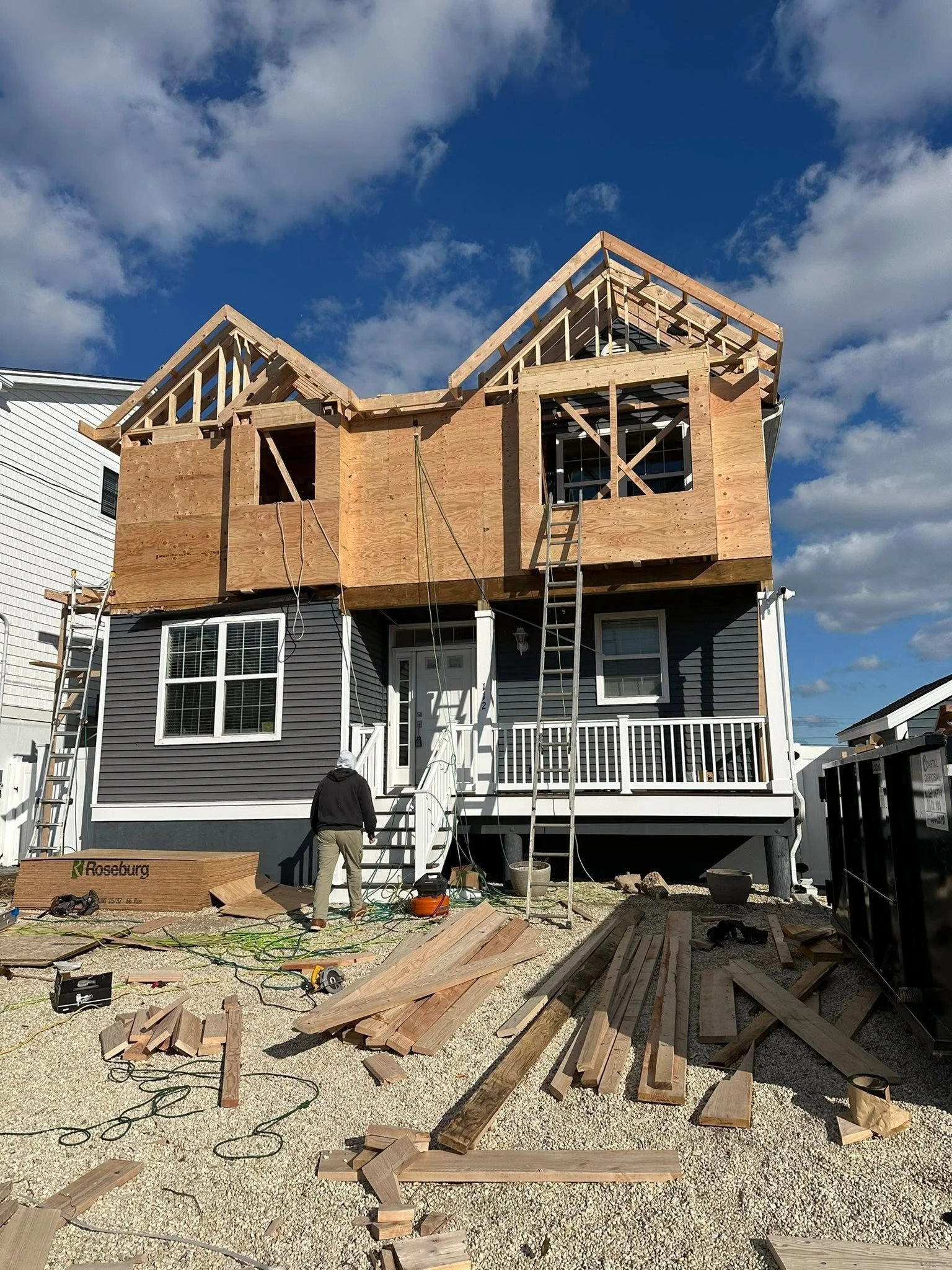 A two-story house under construction with wooden framing. The house has a gray exterior with white trim, a porch, and a door. Construction tools and wooden planks are scattered on the ground. Two workers are present, one in a black hoodie and khaki p