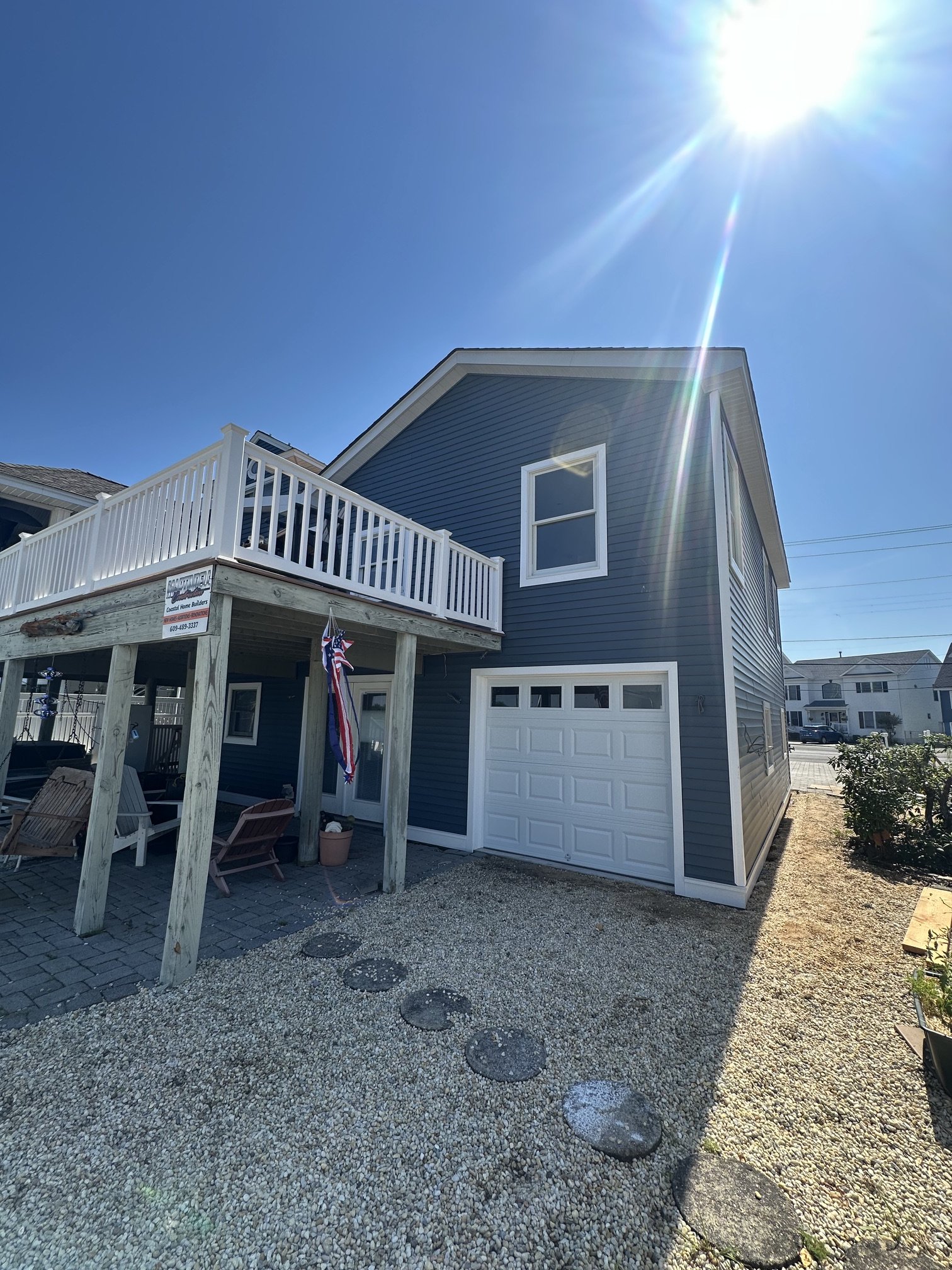 Backyard with gravel ground, stepping stones, and a blue house with a white garage door. There is a porch with a white railing, chairs, and a flag. The sky is bright and sunny.