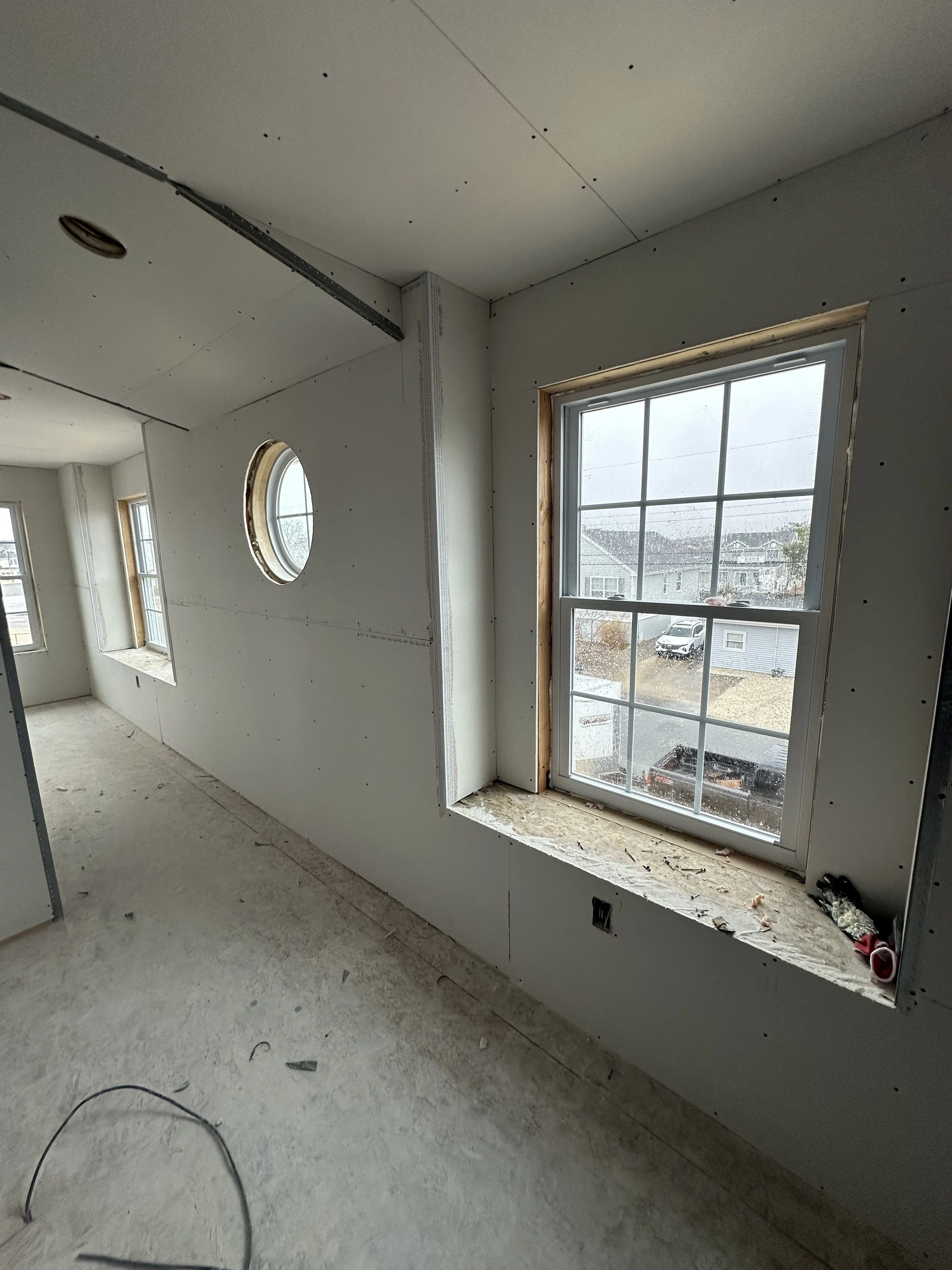 Interior view of a house under construction, showing drywall installation around a window and a circular opening in the wall, with construction tools and debris on the floor.