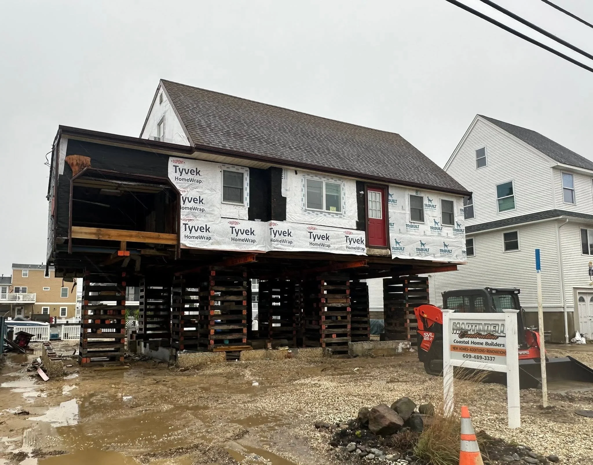 A house under construction on stilts with plywood framing, insulation, and windows, situated at a construction site with muddy ground and construction equipment nearby.