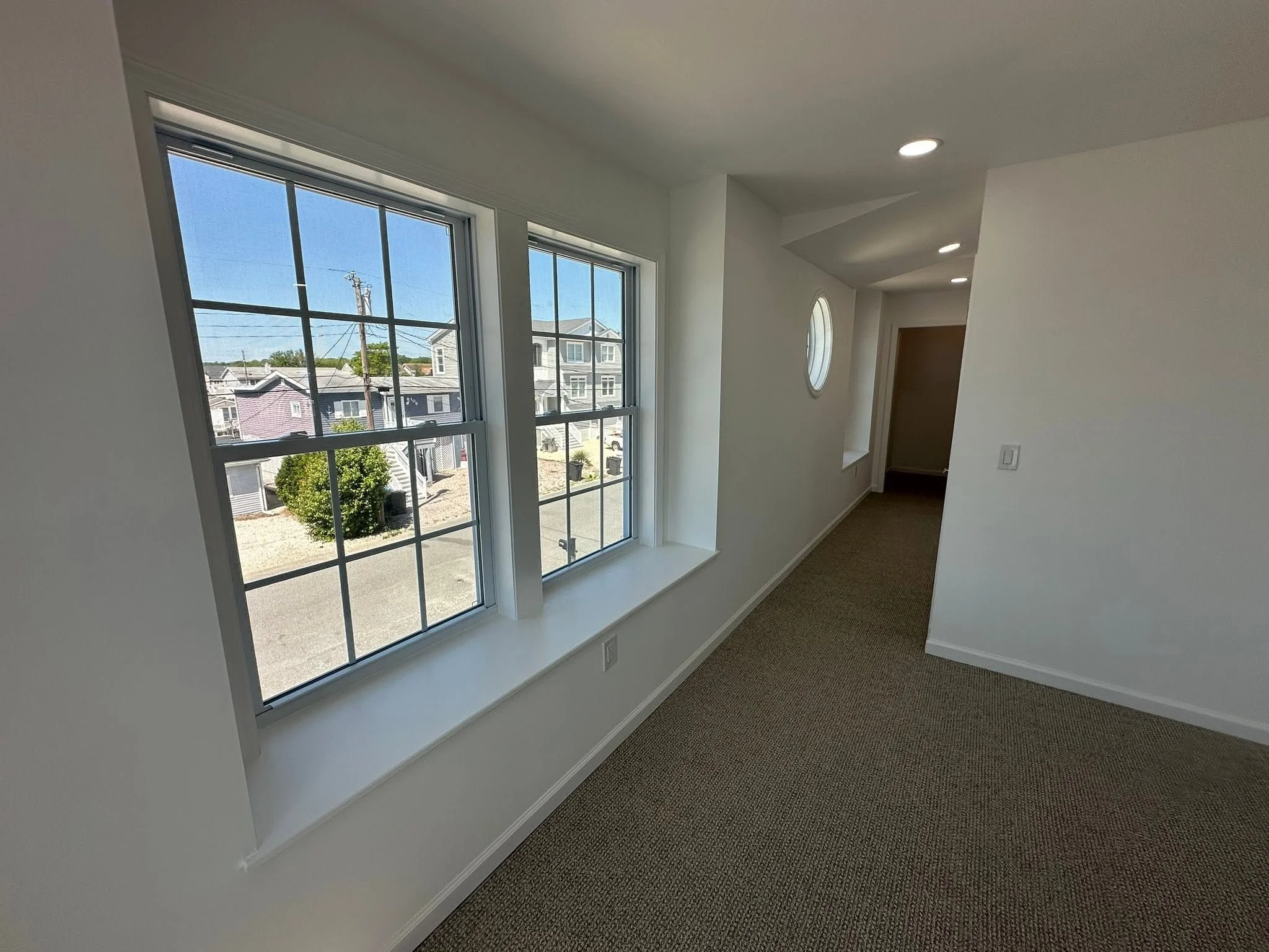 Interior view of a room with white walls, large windows, and ceiling lights. There's a small round window in the wall and a door at the end of the hallway. The floor is carpeted.