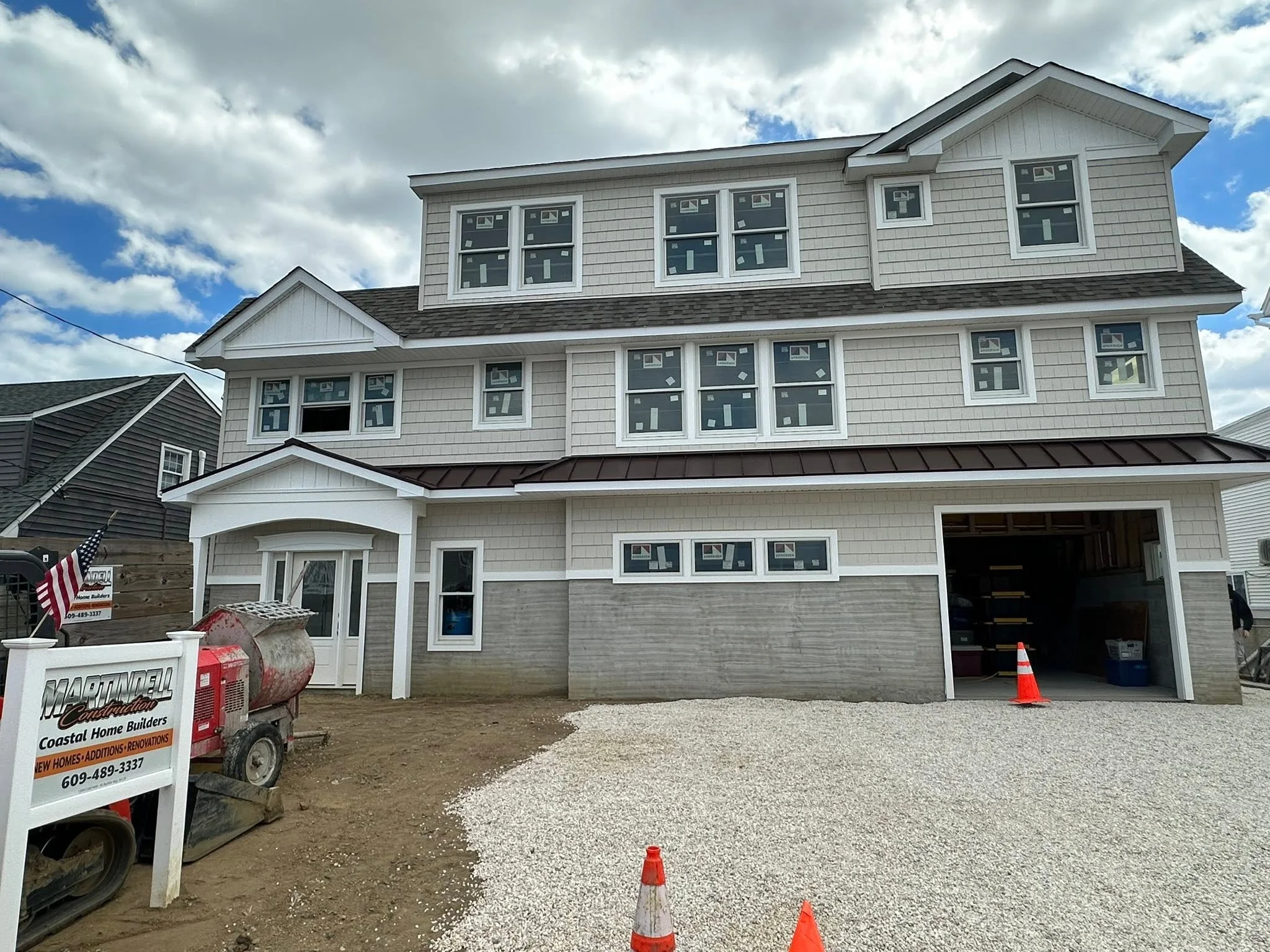 New three-story house under construction with siding and multiple windows, construction equipment, and orange safety cones in the front yard.