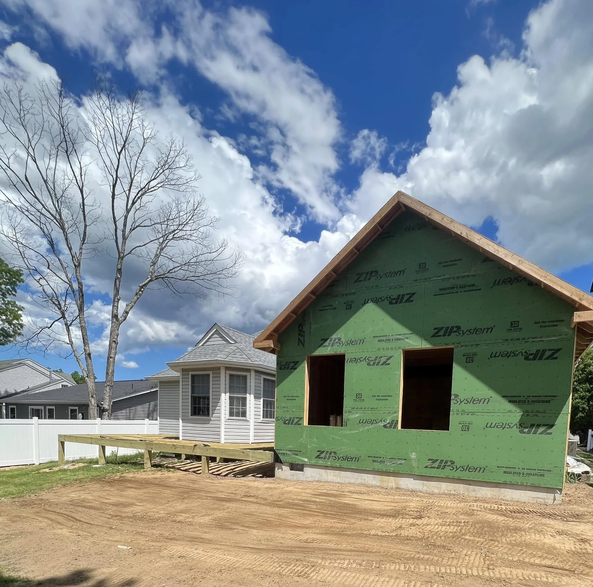 A house under construction with green sheathing, a roof frame, a large bare tree, neighboring finished houses, and a cloudy blue sky.