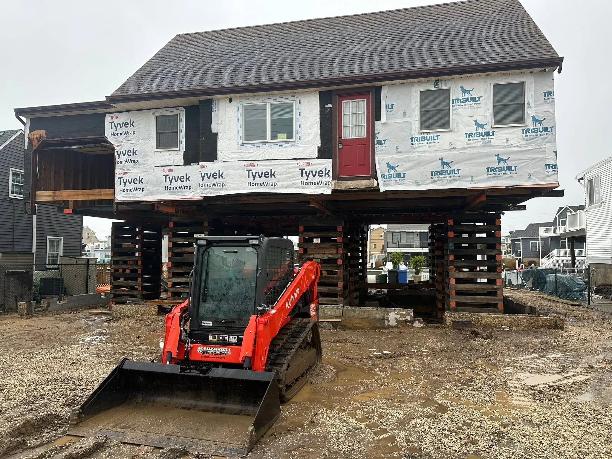 Under construction house with black and white siding, a red door, and a raised foundation. Construction equipment and dirt in the foreground, neighboring houses in the background.