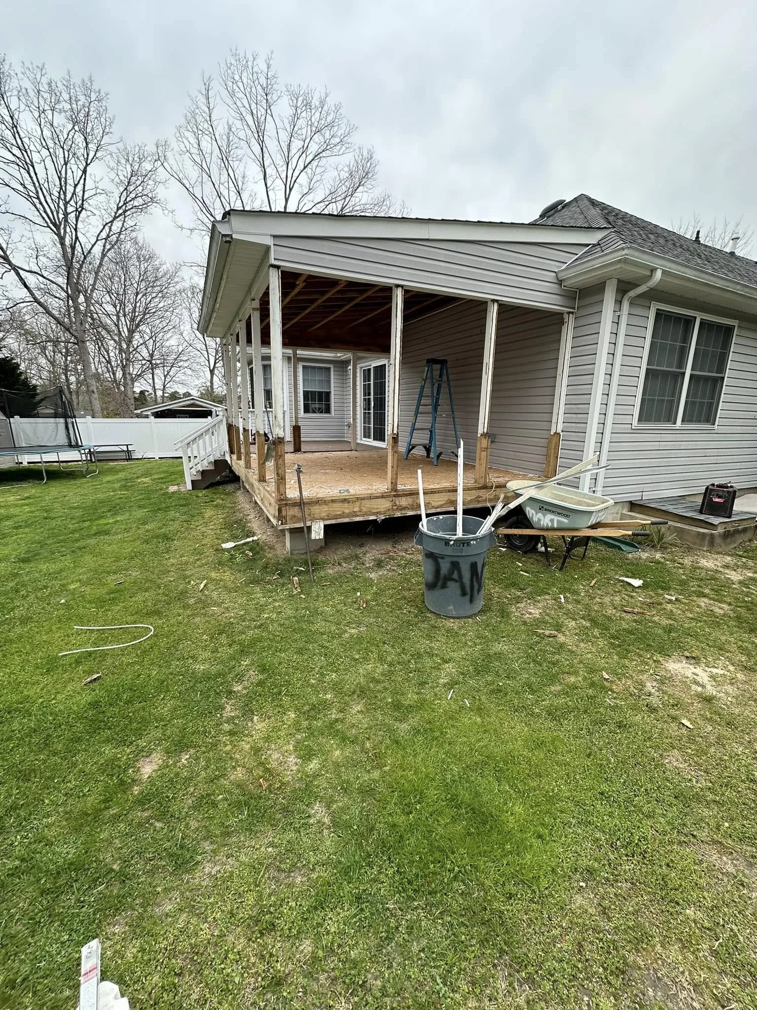 Photo of a house with a partially constructed wooden deck with support posts. There are tools and materials on the ground, including a ladder, a trash bin labeled 'JUNK', and other construction items. The yard is grassy, and there are trees and a whi
