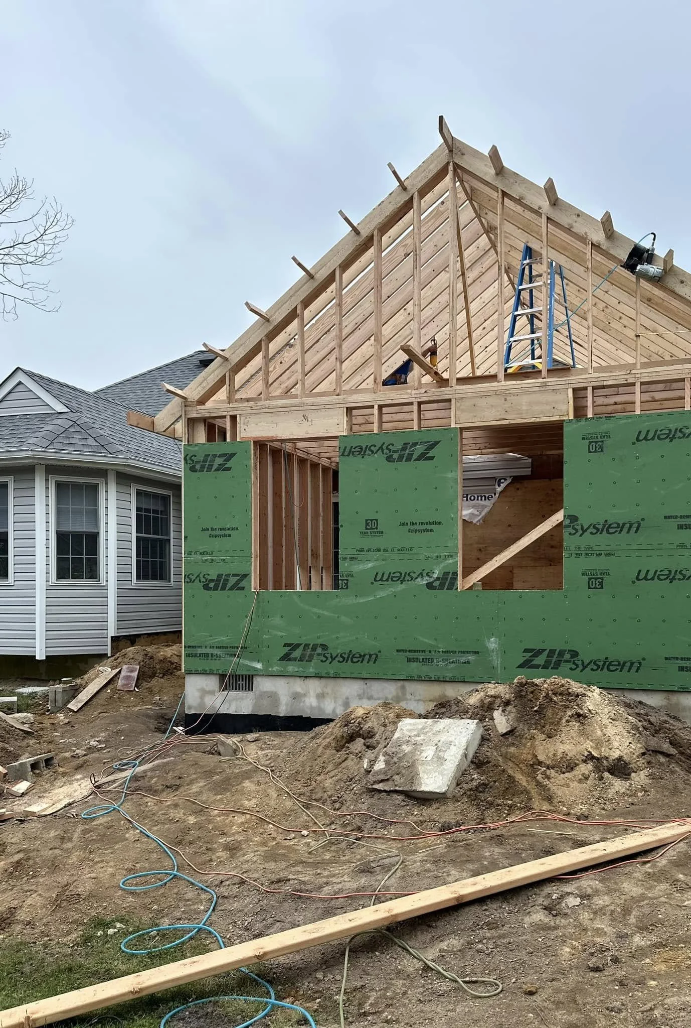 Construction site with a partially built house frame, green insulation sheathing, next to a finished house with gray siding, construction tools, and dirt ground.