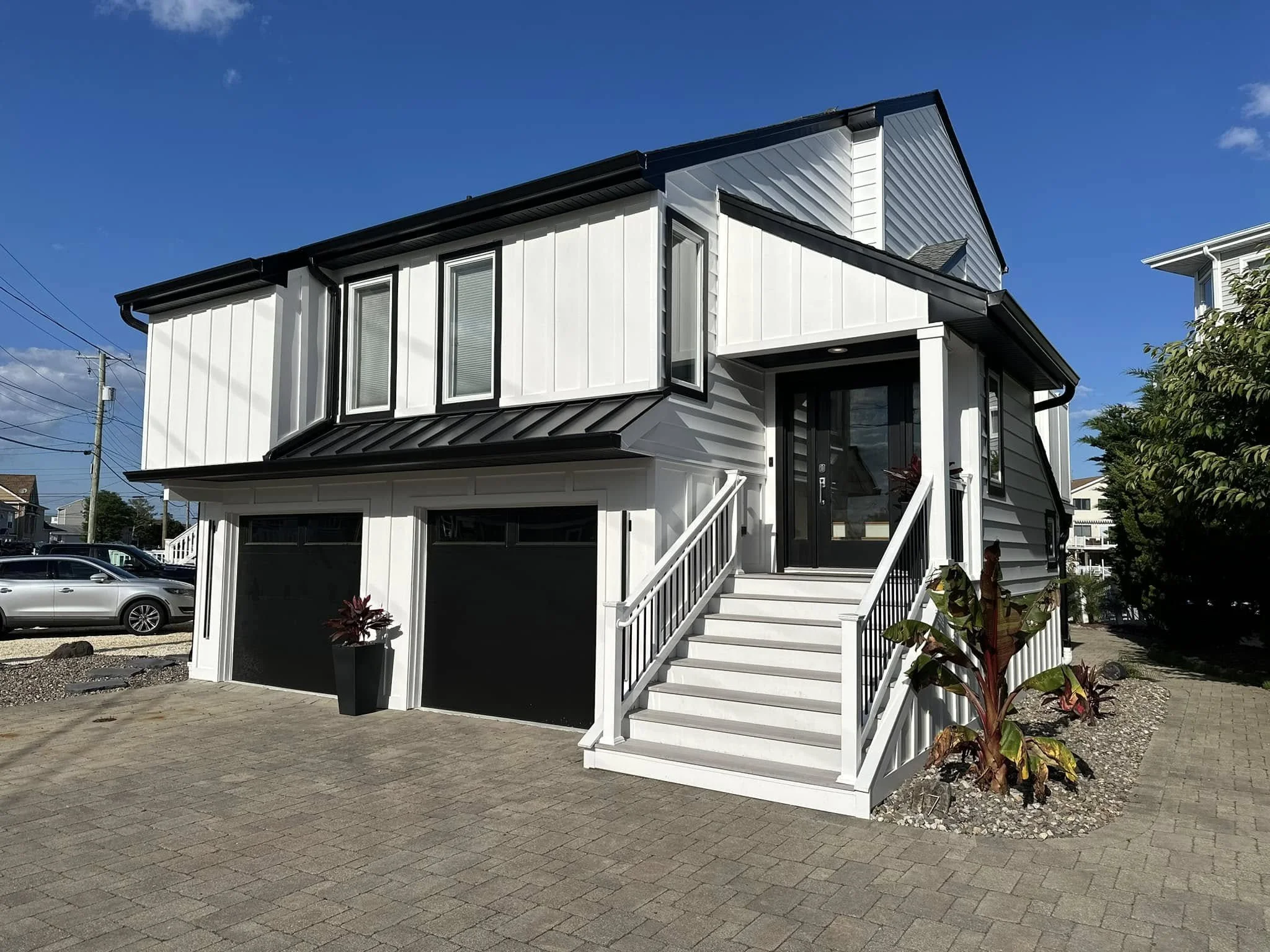 A modern two-story house with white siding and black trim, a staircase with white railings leading to the front door, three narrow windows on the upper level, a garage below, and landscaping with potted plants near the entrance. Blue sky with a few clouds in the background.