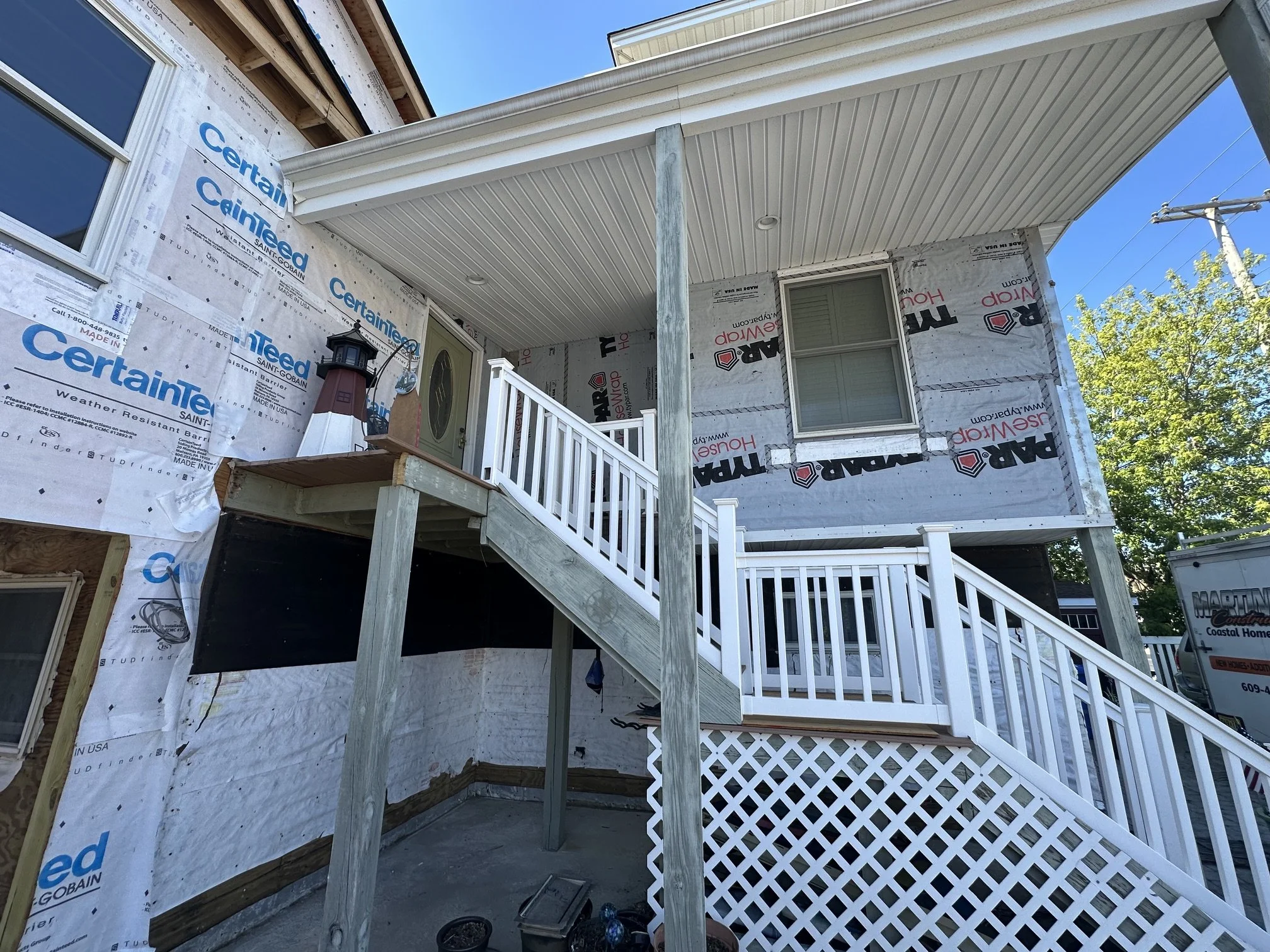 Front of a house under construction with partially finished exterior walls wrapped in weather-resistant barrier, a white staircase leading to a door, and a porch ceiling with recessed lighting. A lighthouse ornament is visible on the porch railing, a