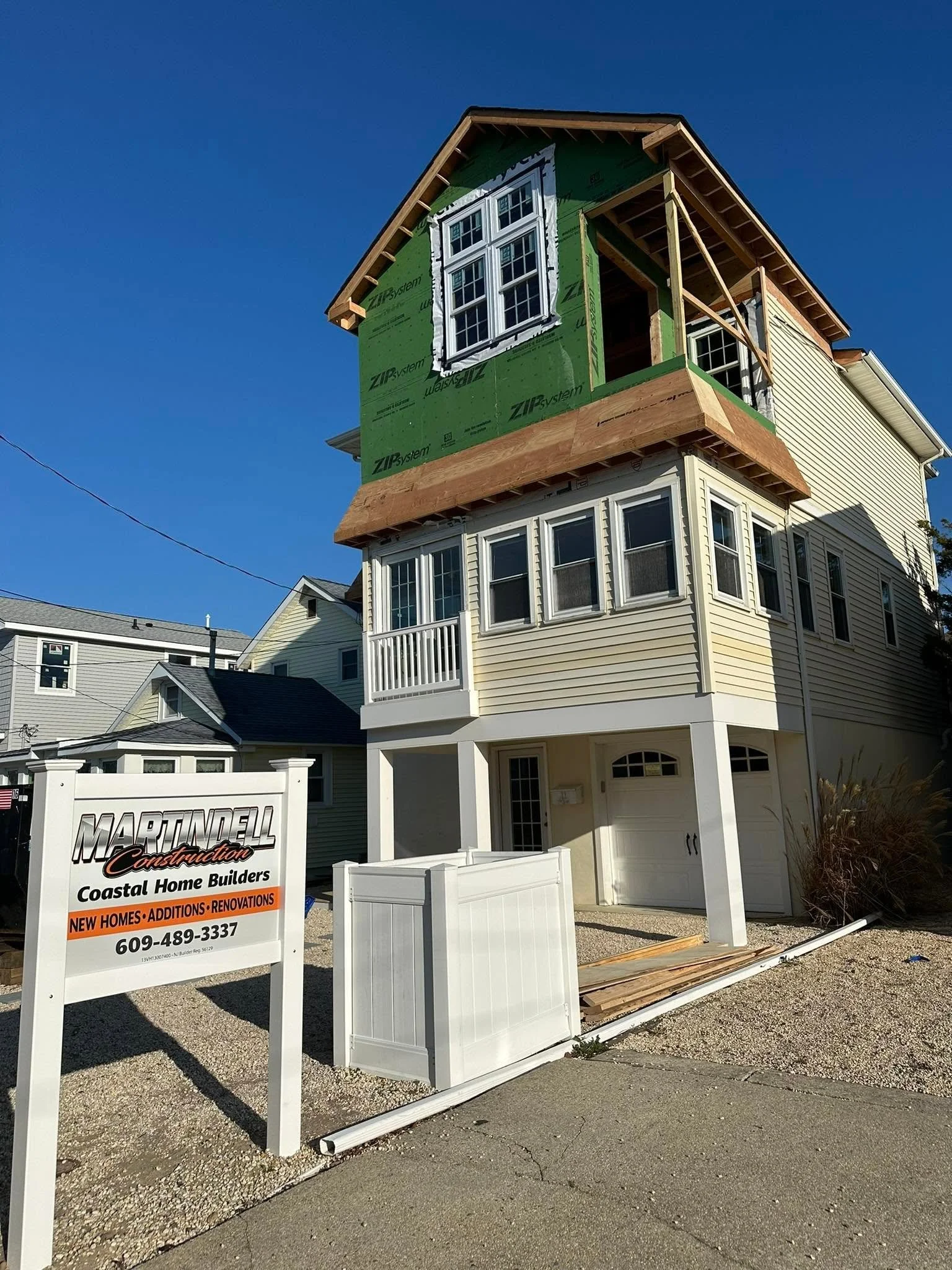 Under construction house with green sheathing, window openings, and siding being installed, construction sign in foreground, blue sky overhead.