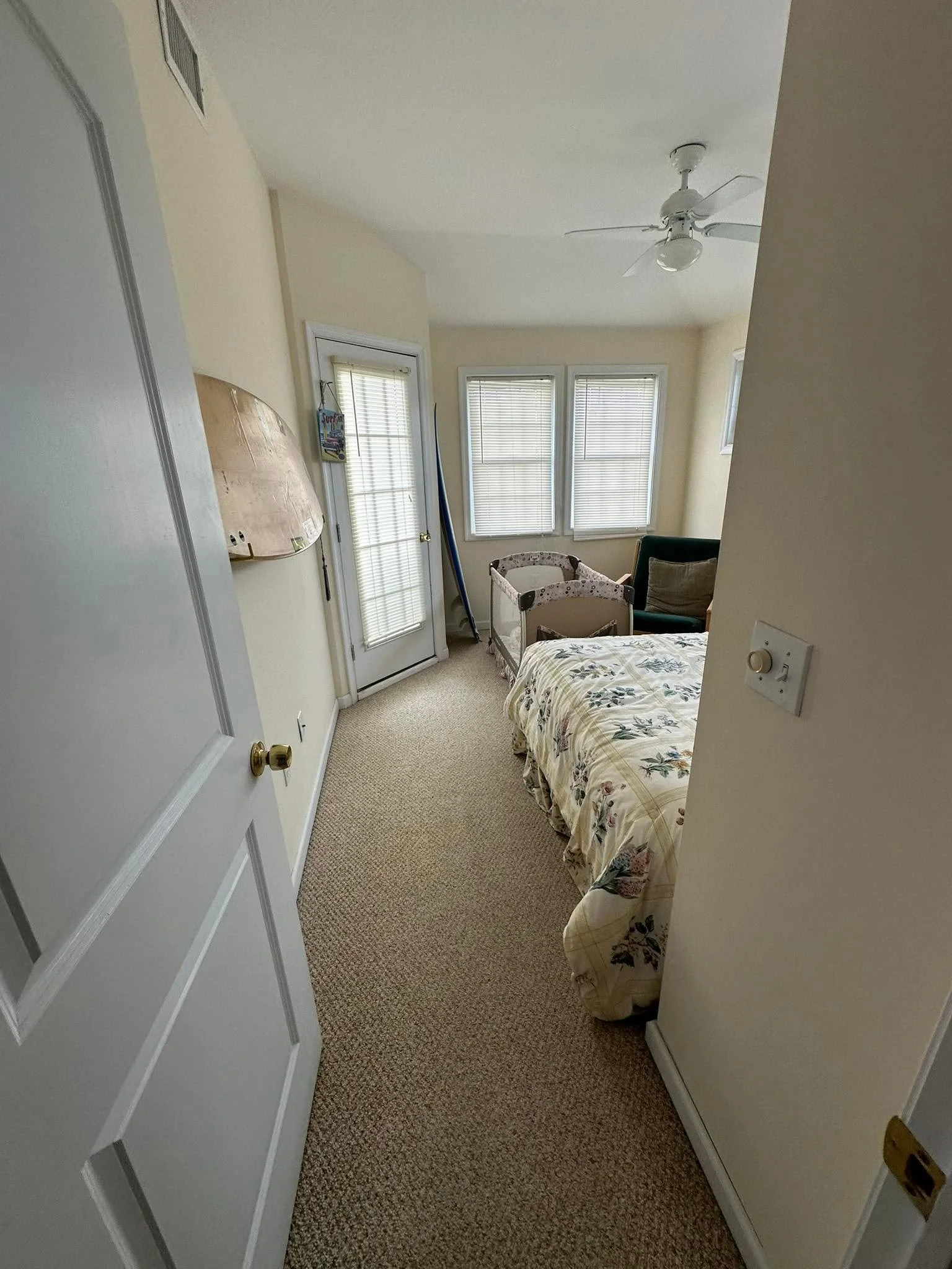 A small bedroom with a bed covered in floral bedding, a green armchair with a pillow, a crib, and three windows with blinds. There's a ceiling fan and a door with blinds, a mirror, and a hanging decoration.