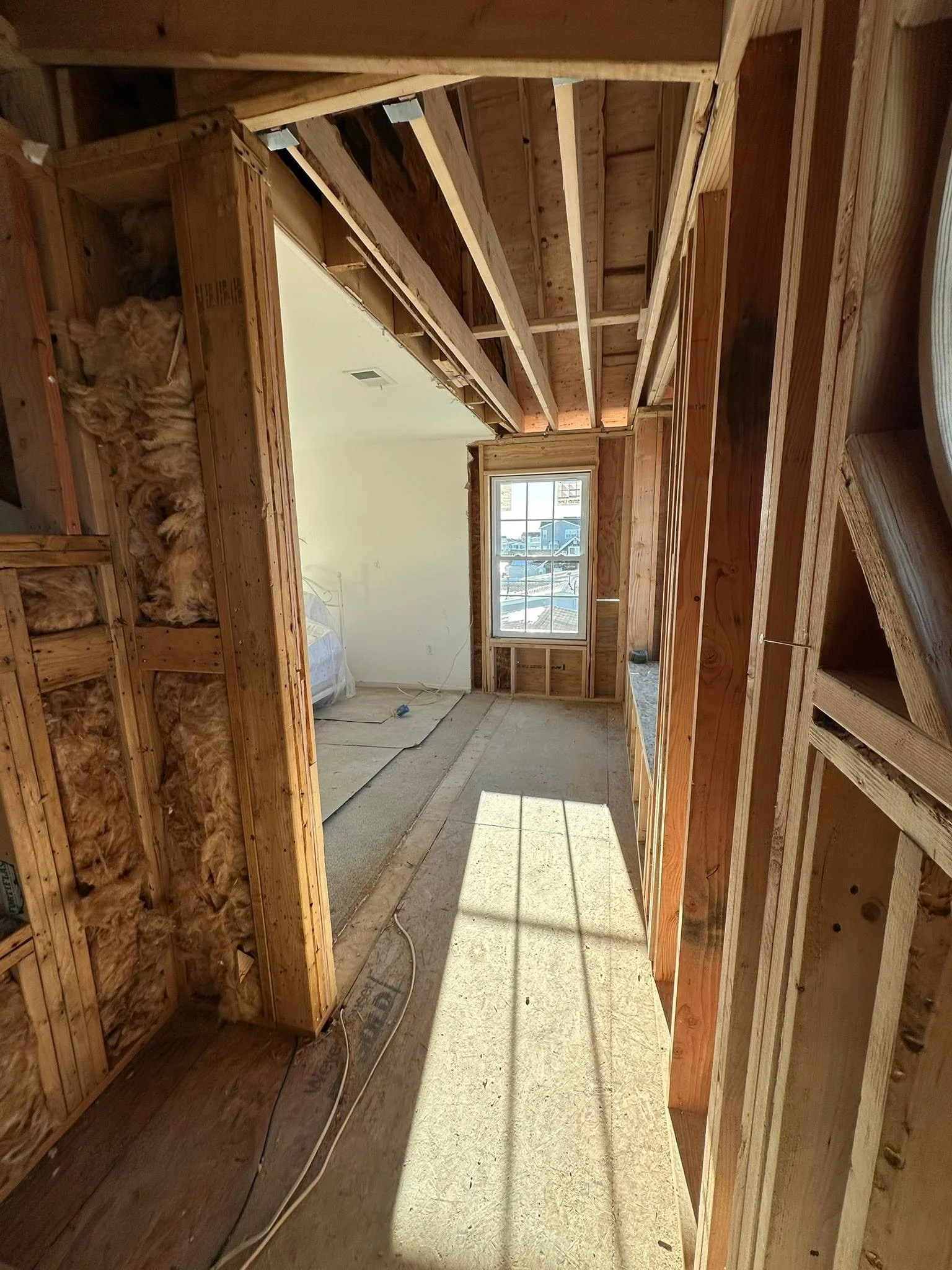 Interior view of a house under construction, showing wooden wall framing, insulation, and an unfinished doorway with a window in the background.