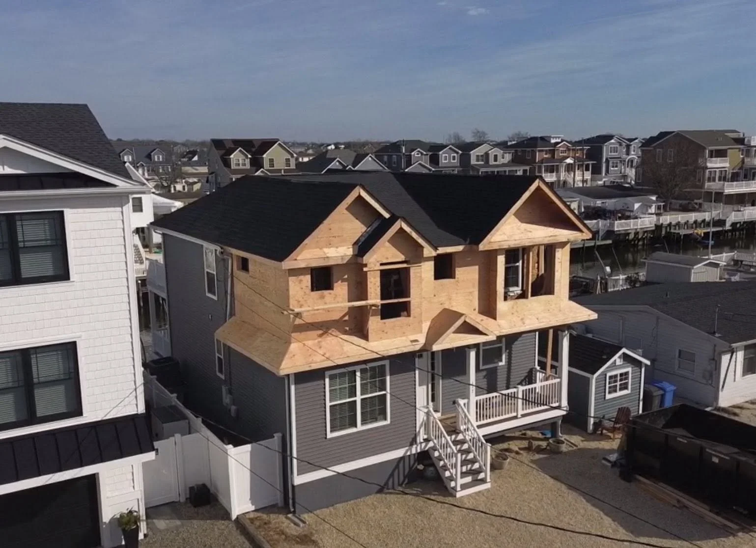 A house under construction with wooden framing and a dark shingled roof, located in a neighborhood with other completed houses and docks along a waterway.